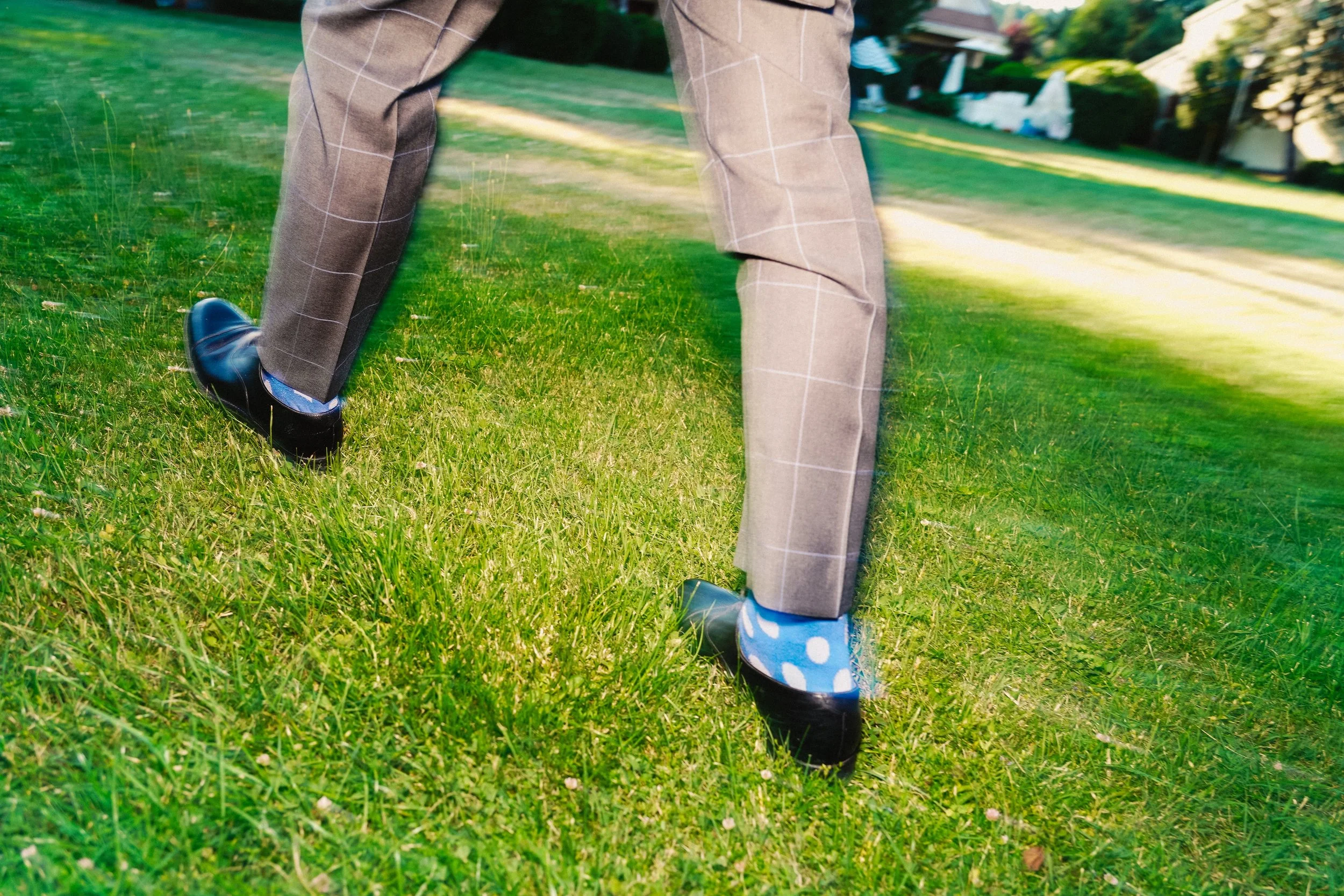 Persona usando zapatos negros, con calcetines azules con puntos blancos, caminando o ajustando el pantalón en un césped en un entorno residencial.