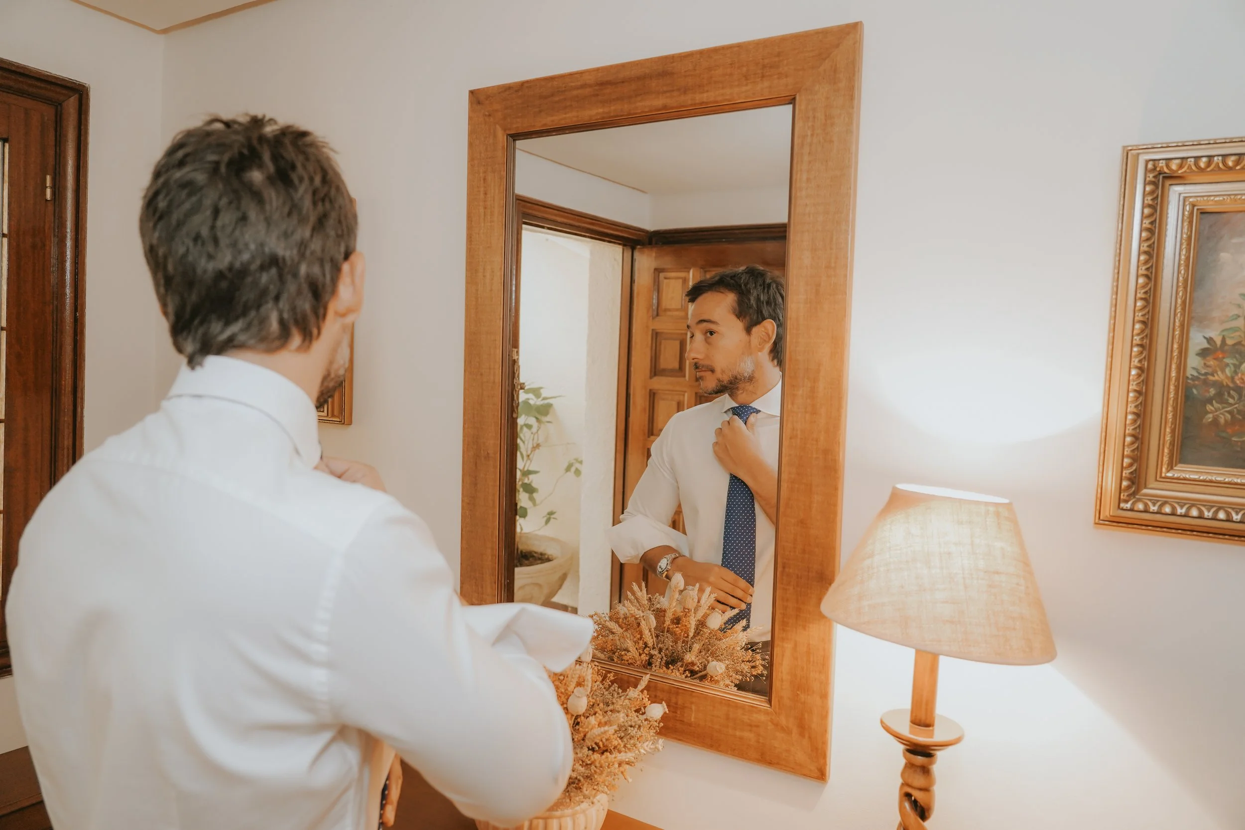 Un hombre con camisa blanca y corbata azul ajustándose en un espejo, mientras otro hombre, también con camisa blanca, lo observa desde atrás en una habitación decorada con cuadros y una lámpara
