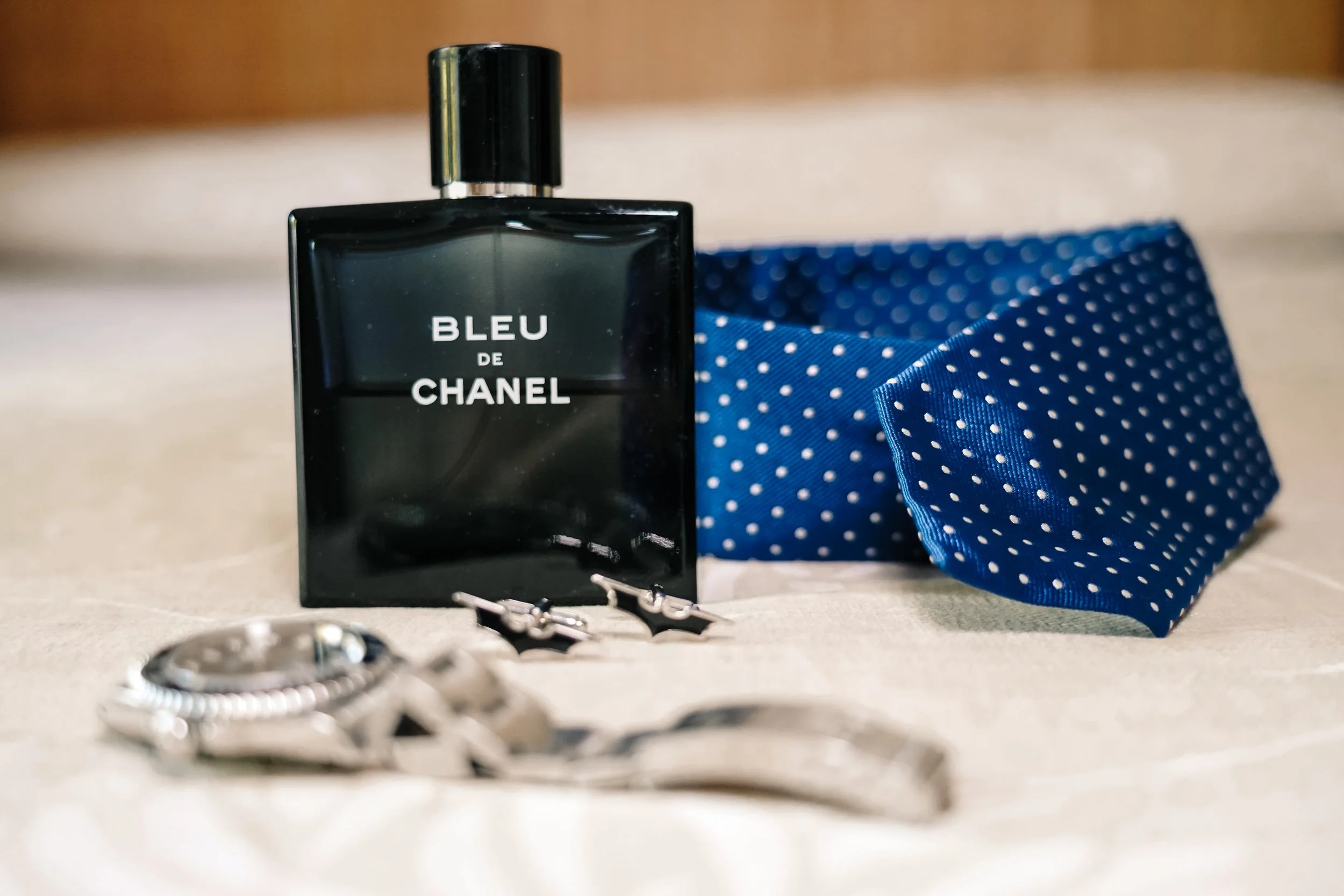 Botella de perfume Bleu de Chanel, una pajarita azul con puntos blancos, unos pendientes y un reloj de plata, todo sobre una superficie clara.