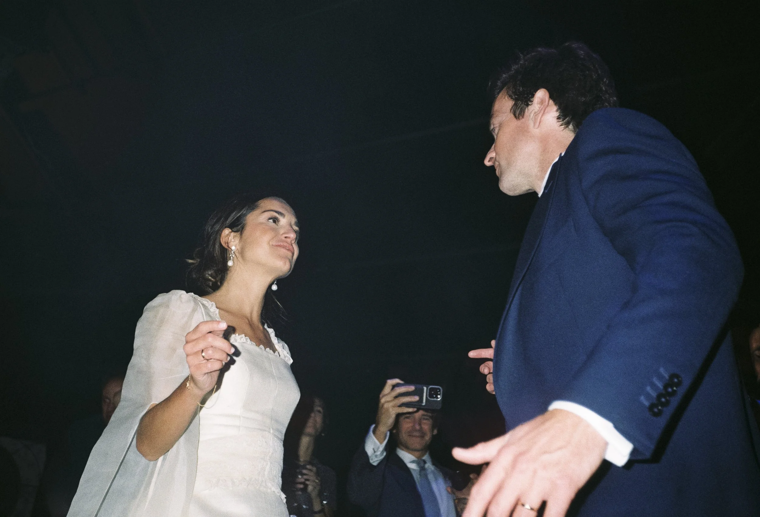 Una mujer y un hombre en una especie de reunión o evento formal, interactuando entre sí. La mujer lleva un vestido blanco y pendientes largos, y el hombre viste un traje azul oscuro. Ella parece estar entregándole un objeto, posiblemente un anillo o algo simbólico, mientras otros en el fondo toman fotos o ven la escena.