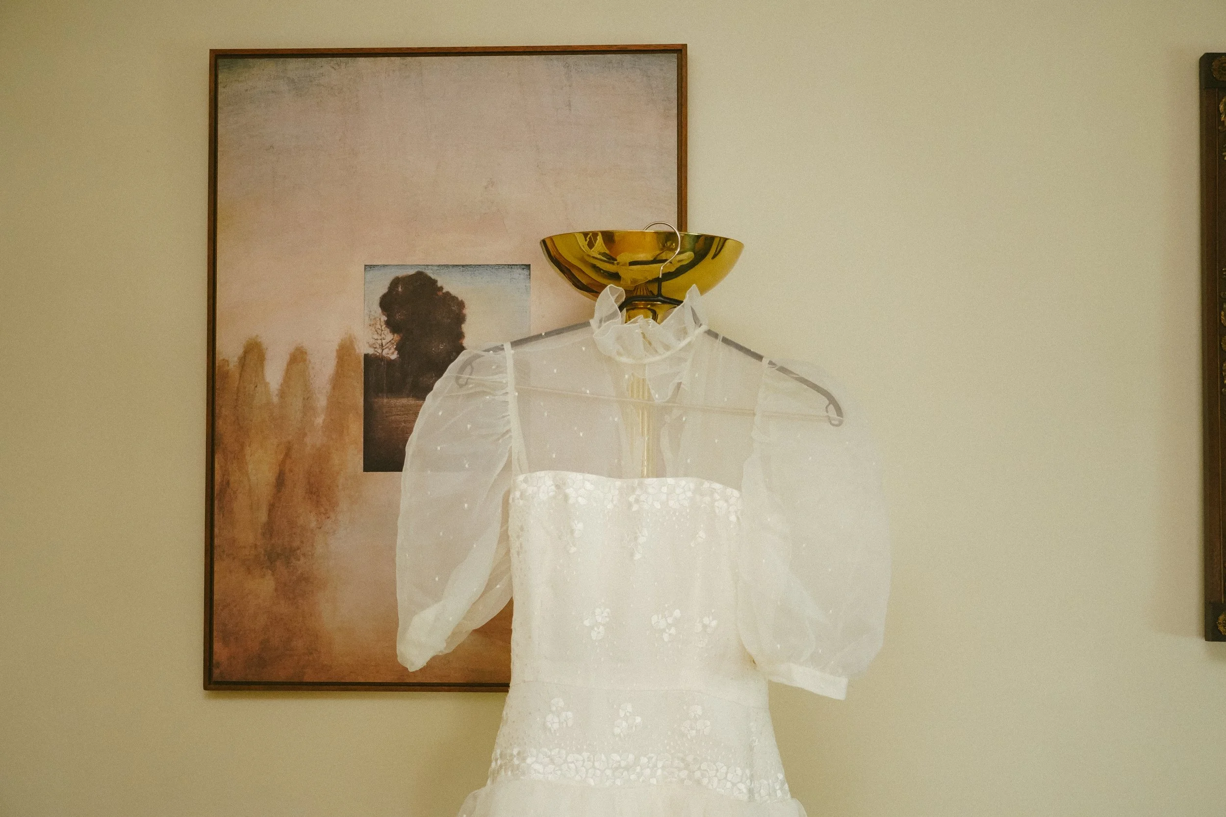 Vestido blanco de encaje con mangas transparentes y lazo en el cuello, colgado sobre un maniquí, con un cuadro y un florero dorado en el fondo en una pared de color claro.