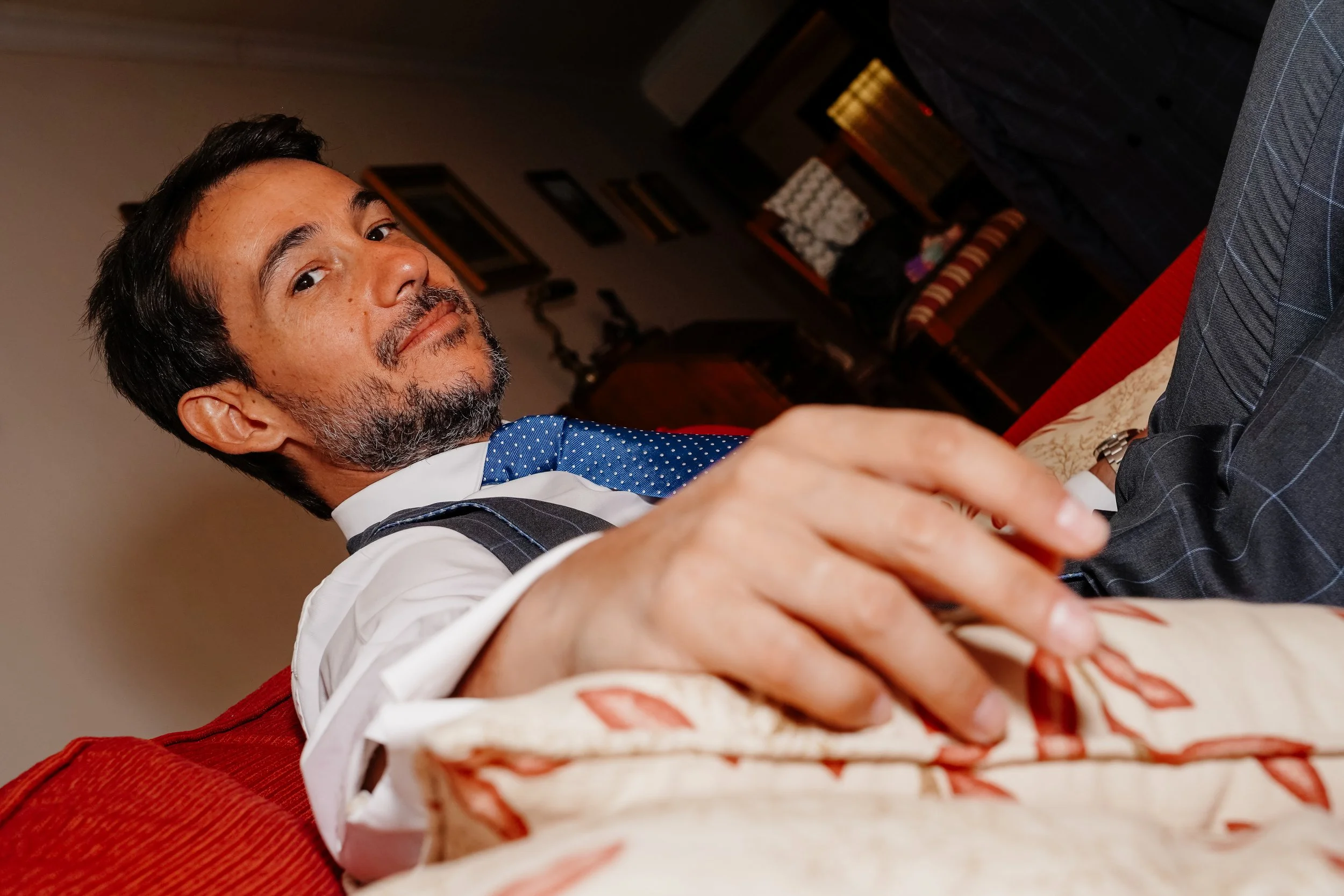 Hombre con barba en traje y corbata sentándose en un sofá, mirando hacia la cámara.