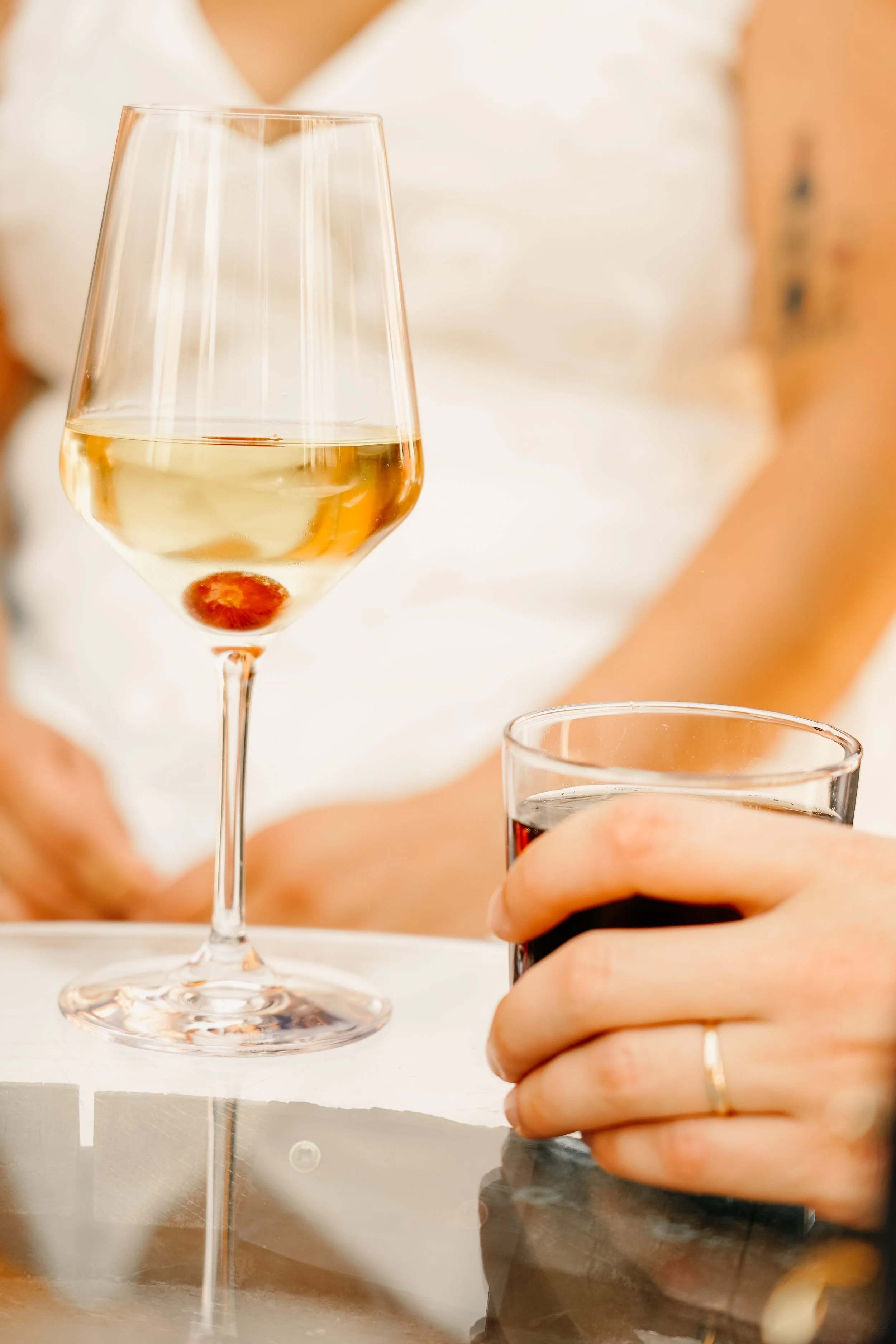 Persona sosteniendo una copa de vino blanco y un vaso de refresco oscuro en una mesa de cristal.