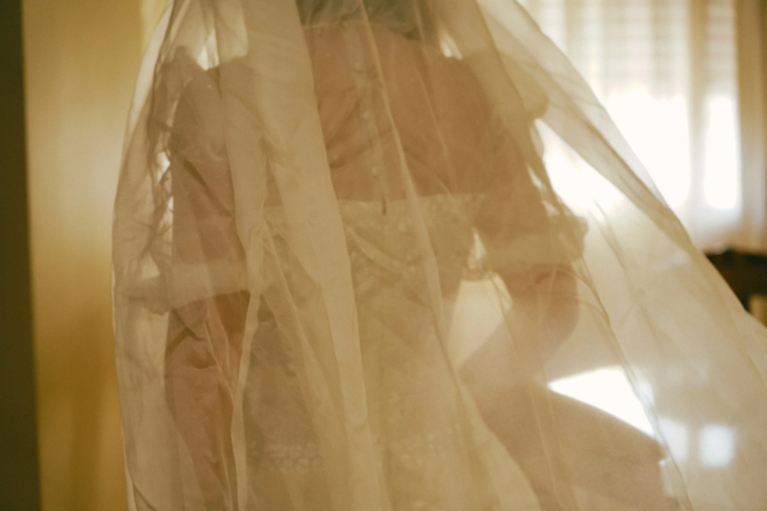 Una persona con un vestido largo y transparente, parcialmente oculta detrás de un velo, en un ambiente interior con luz suave y cortinas blancas.