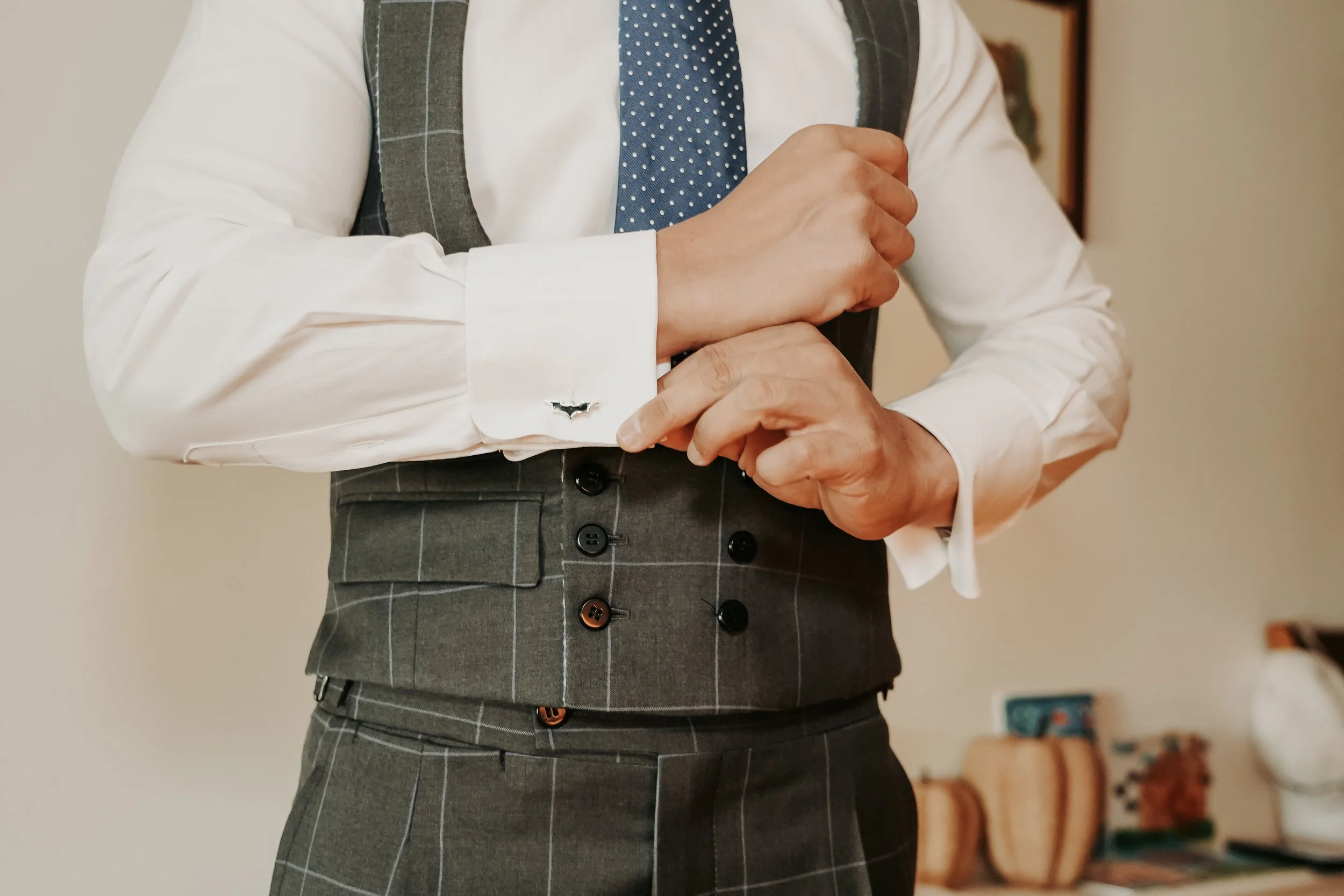 Hombre con camisa blanca, chaleco gris con línea de cuadros y corbata azul, ajustándose la manga de la camisa en un ambiente interior.