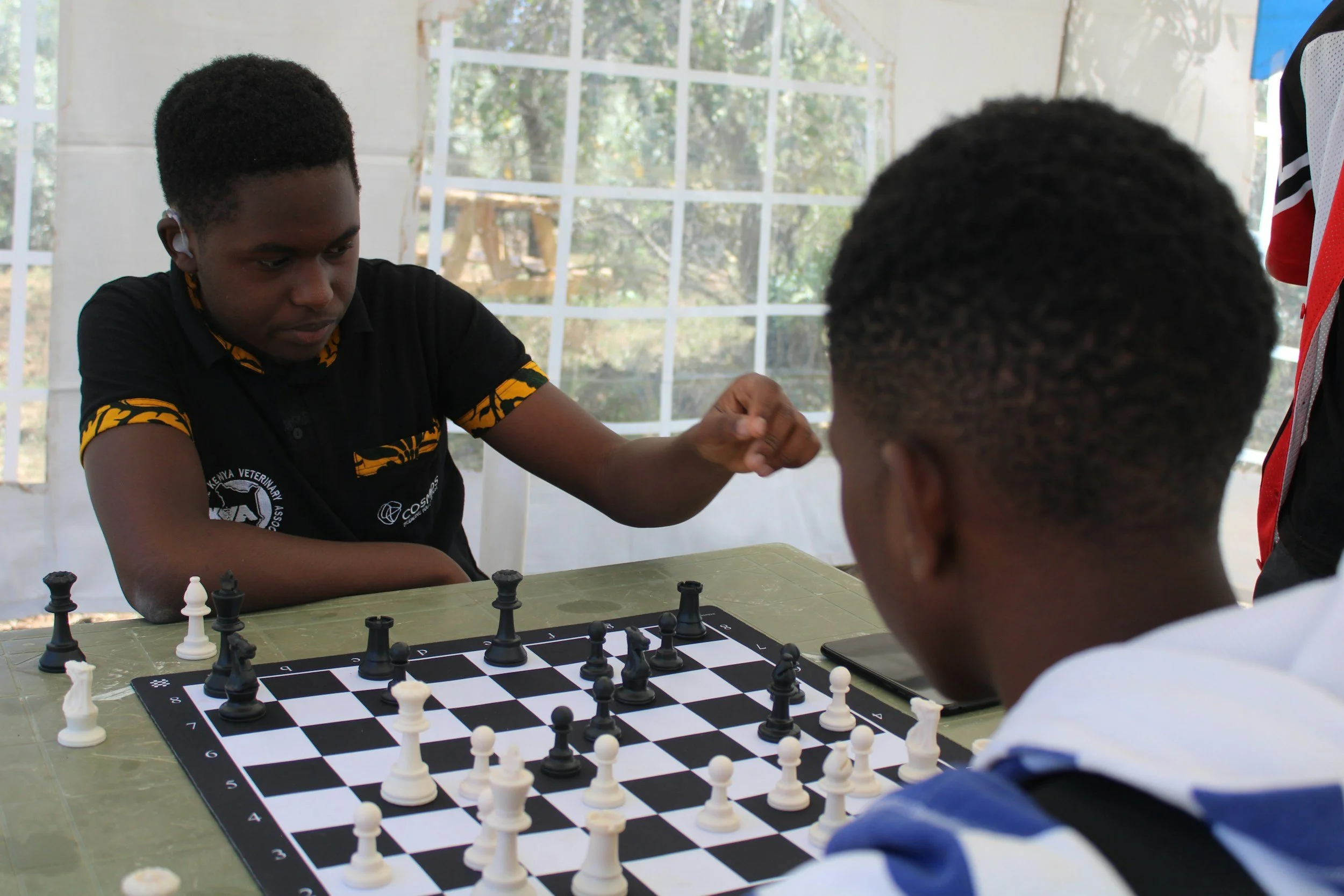 Two young people playing chess indoors, with one making a move and the other observing, on a green table in front of a large window