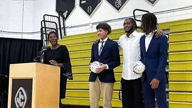 Four people standing on yellow bleachers, with one at a podium and three holding rugby balls.