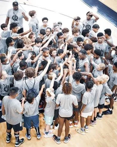 A large group of children and a few adults gathered on a basketball court, participating in a coordinated activity or game, all wearing gray T-shirts.