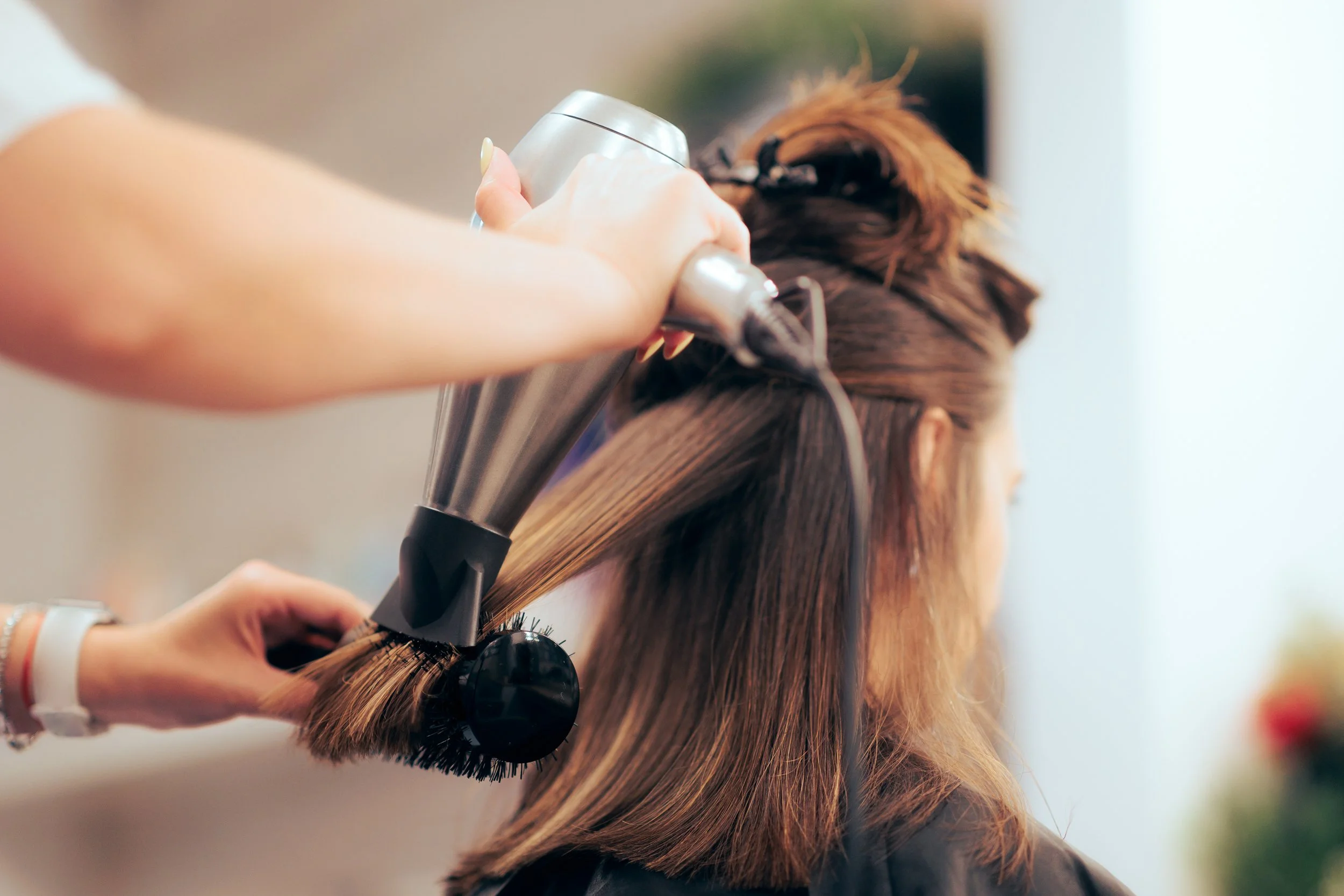 A hairstylist blow-drying a woman's brown hair in a salon.
