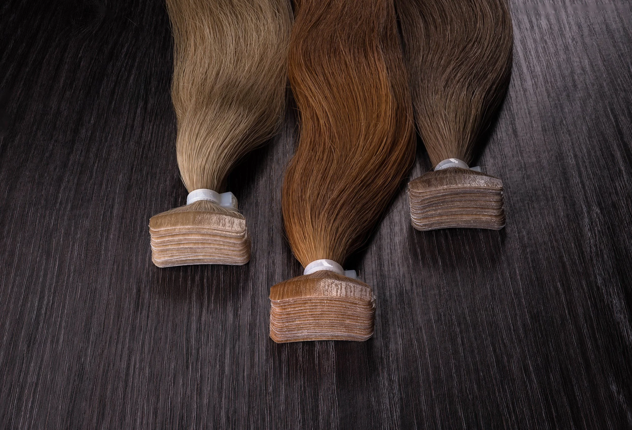 Three bundles of hair extensions in shades of blonde, red, and brown, arranged on a dark wooden surface.