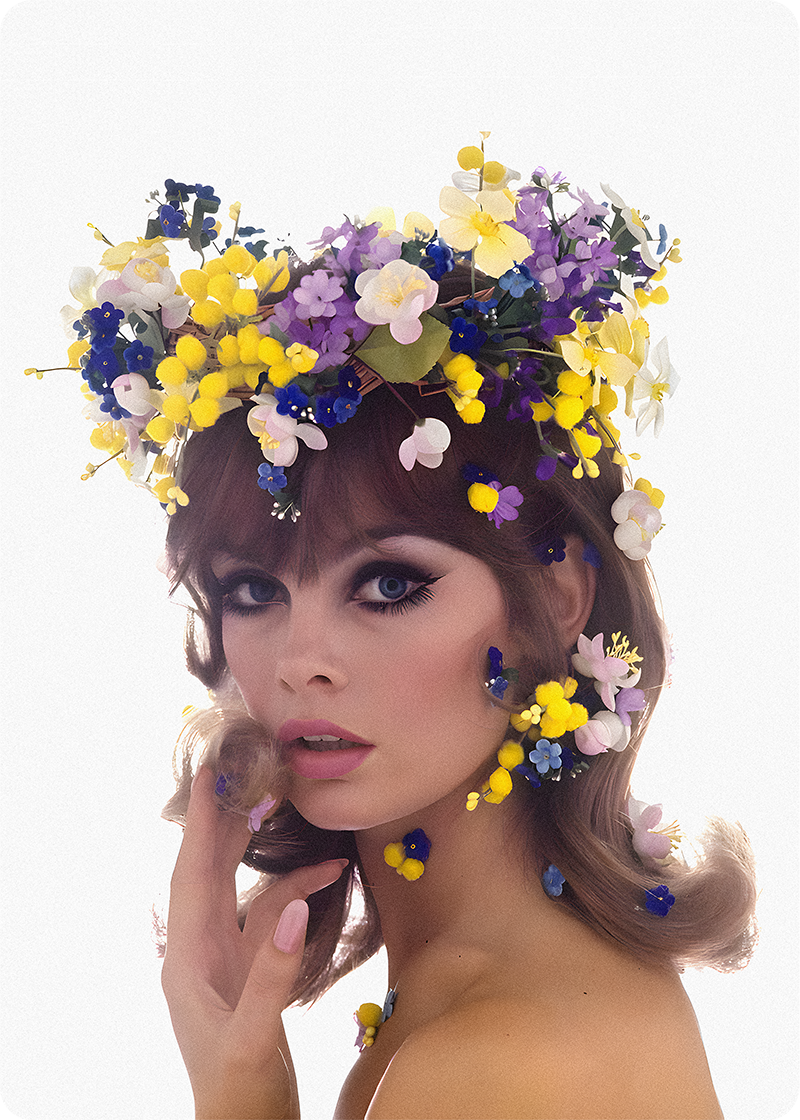 Woman with a floral crown made of yellow, purple, white, and blue flowers, with some flowers on her face and shoulder, posing against a plain white background.