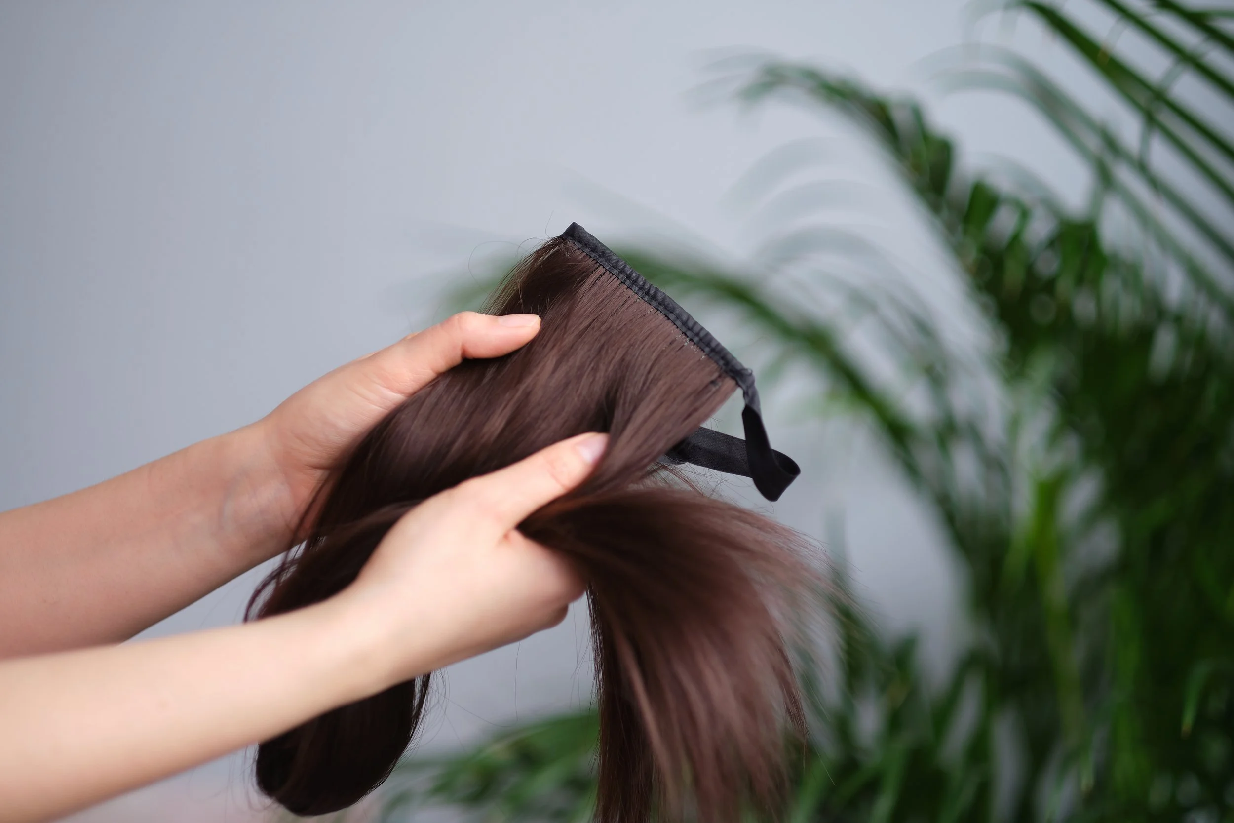 Person holding a brown hair extension or wig with a black lace closure against a grey background and green plants.
