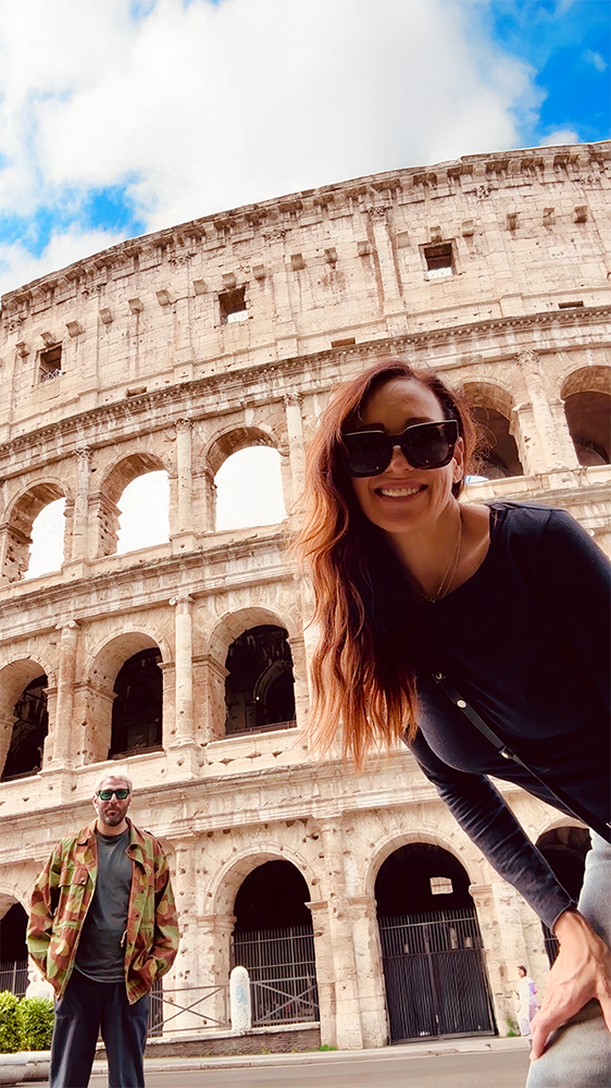 Smiling woman with long red hair and large sunglasses taking a selfie in front of the Colosseum in Rome, Italy, with a man standing in the background.