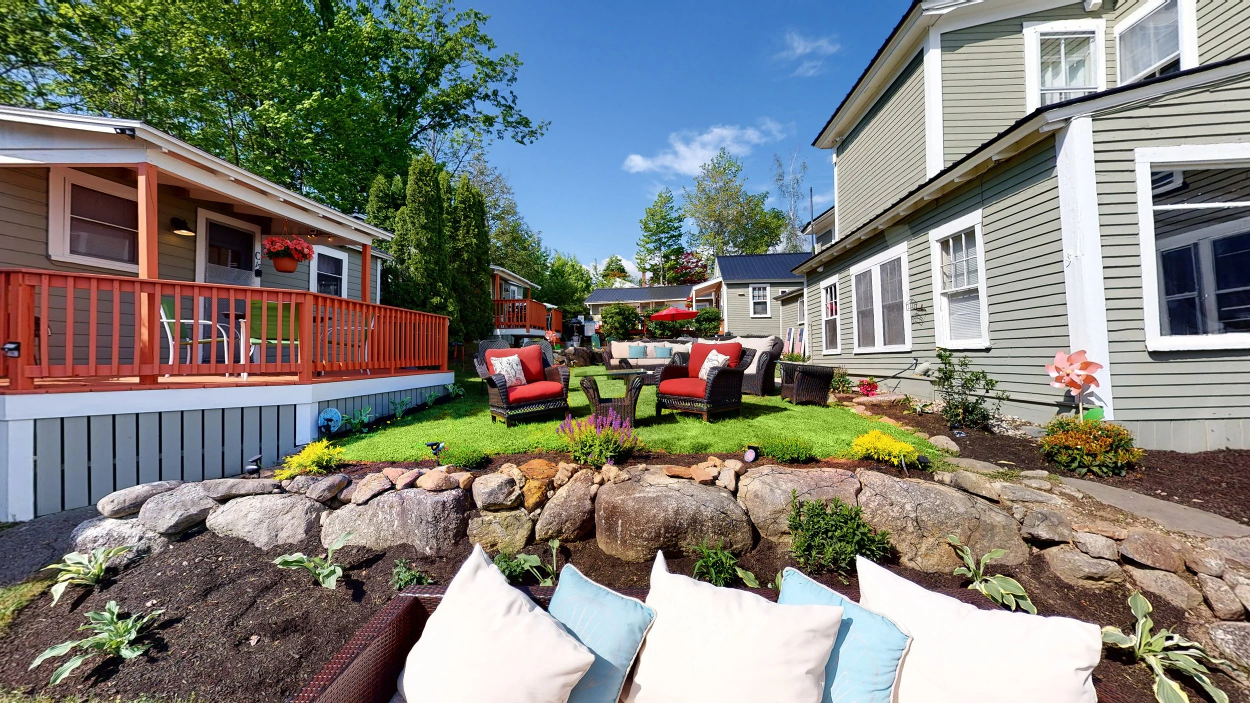 Outdoor patio area with wicker furniture, red cushions, and surrounding cottages, set in a landscaped garden with stone accents and flowers.