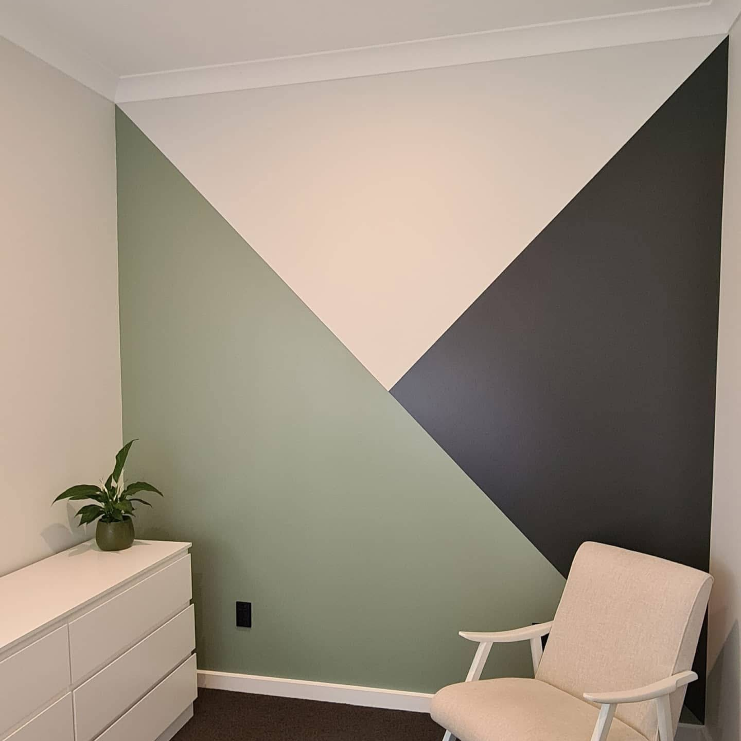 A corner of a room with a geometric painted wall, a white dresser with a plant, and a beige chair.