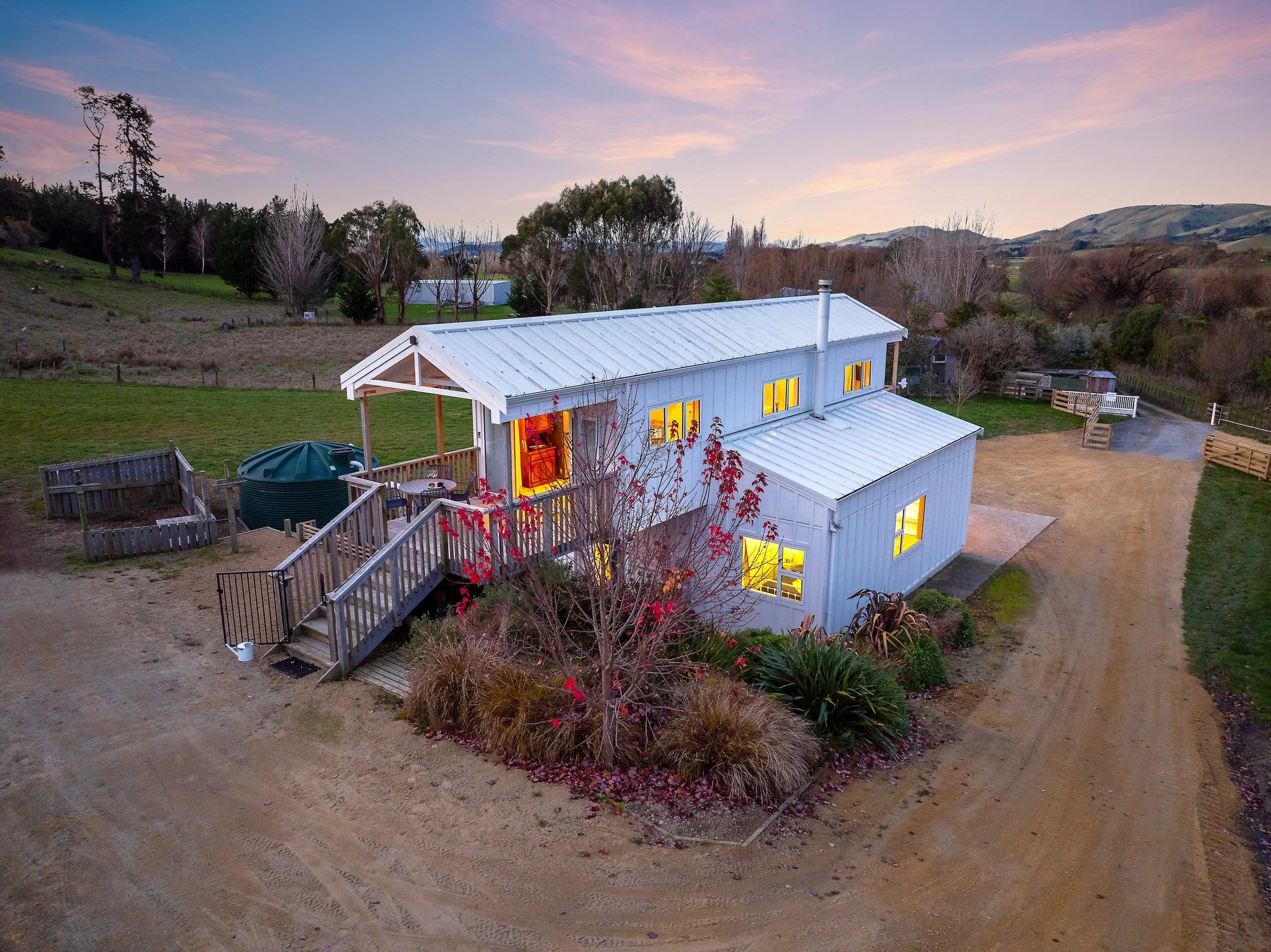 Elevated rear view of barn style white house at dusk with blue/pink sky in background. Lights are on inside.
