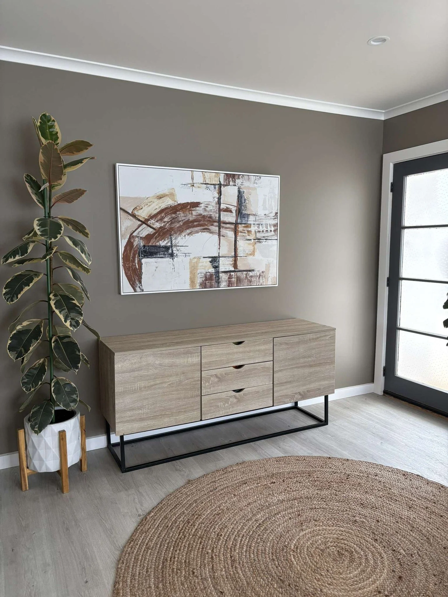 Interior of home painted mushroom grey showing low drawers, modern art, a plant and a round rug. A wooden framed glass door is at the right.