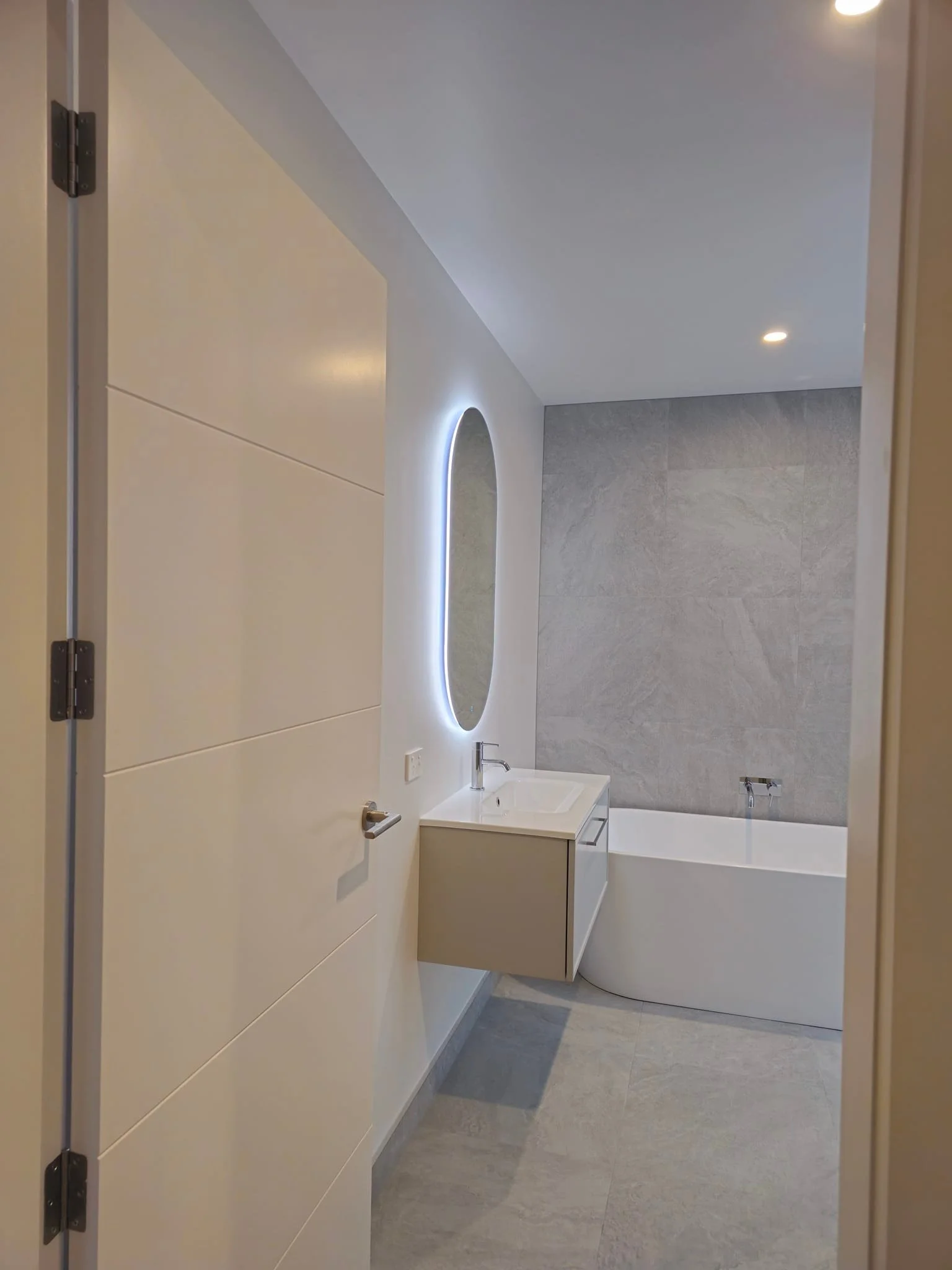 Modern bathroom with a floating vanity, illuminated mirror, bathtub, gray tiled wall, and recessed lighting.
