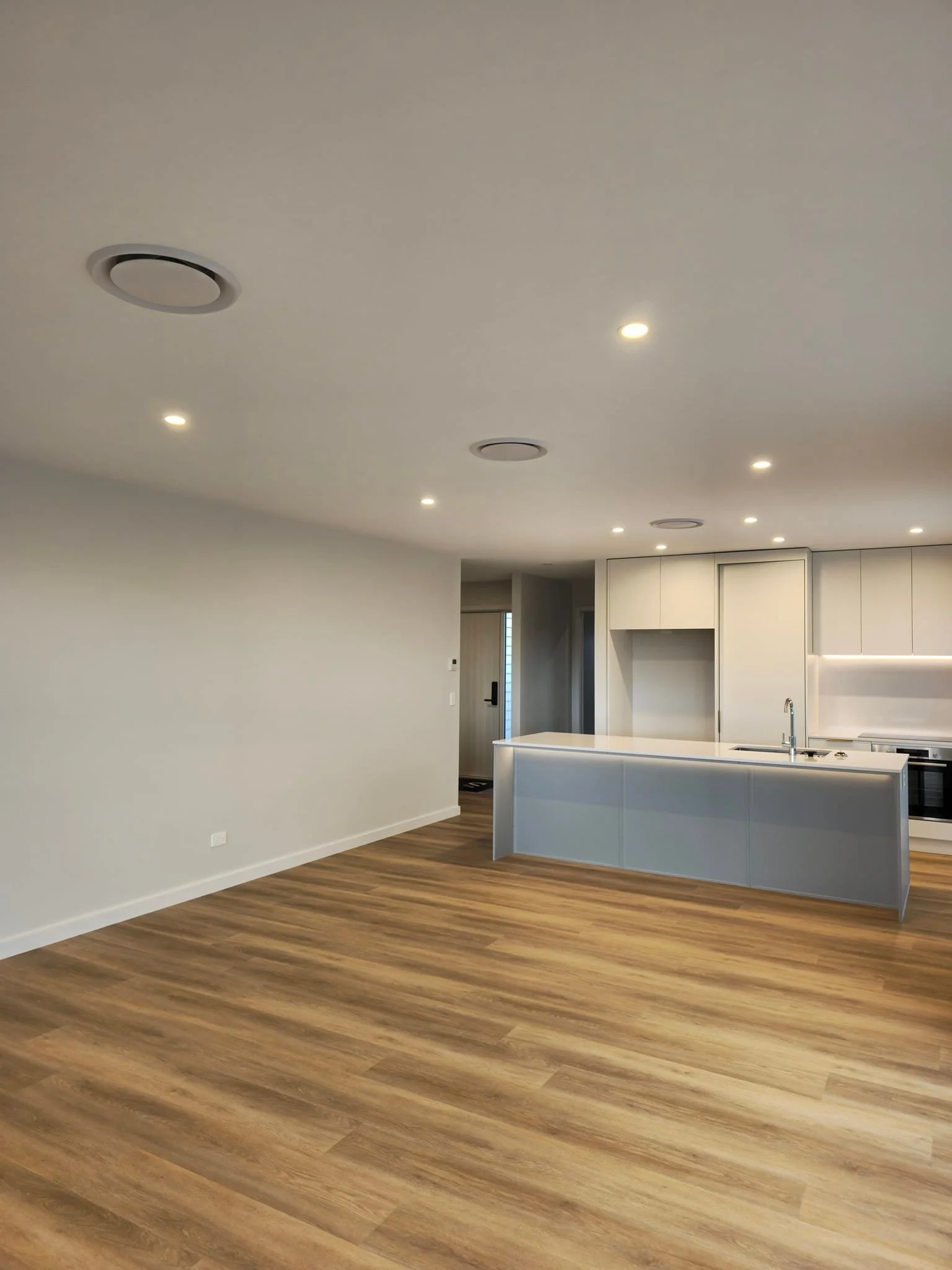 Interior view of a modern, empty living space with wood flooring, white walls, and a kitchen area with white cabinets and a countertop.