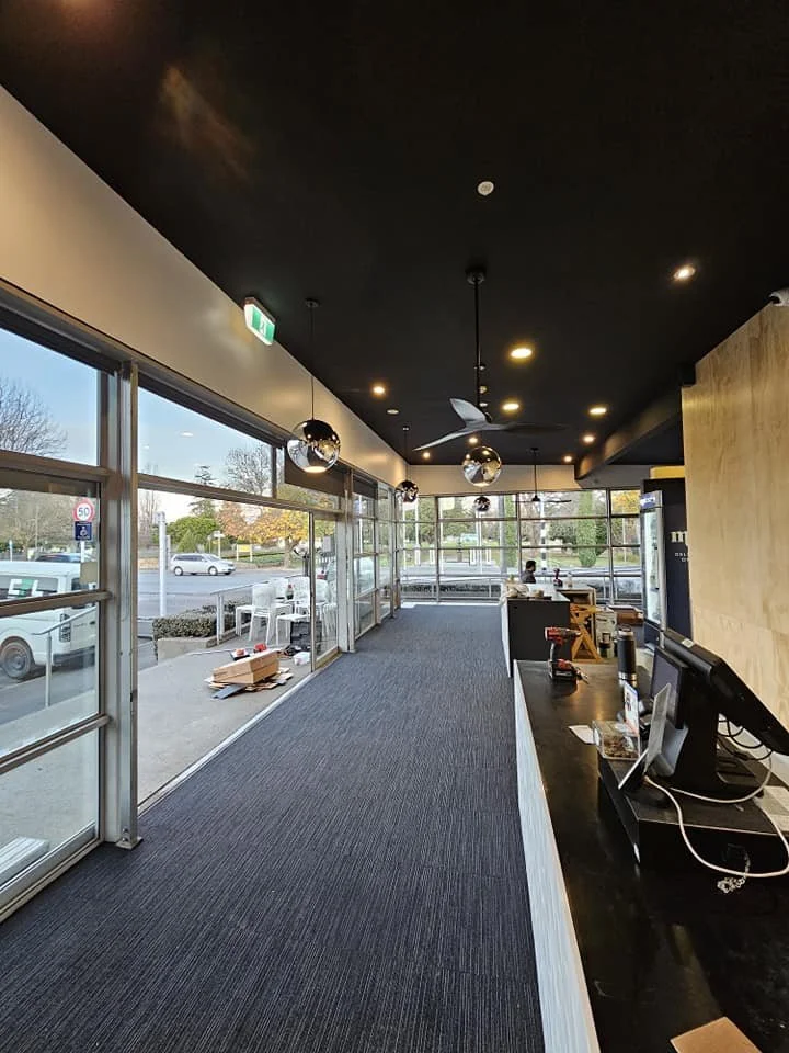 Interior view of a modern cafe with black ceiling, black carpet, wooden wall on the right, large window to the left and at the rear, and hanging reflective spherical lights. There is a black counter with coffee equipment to the right.