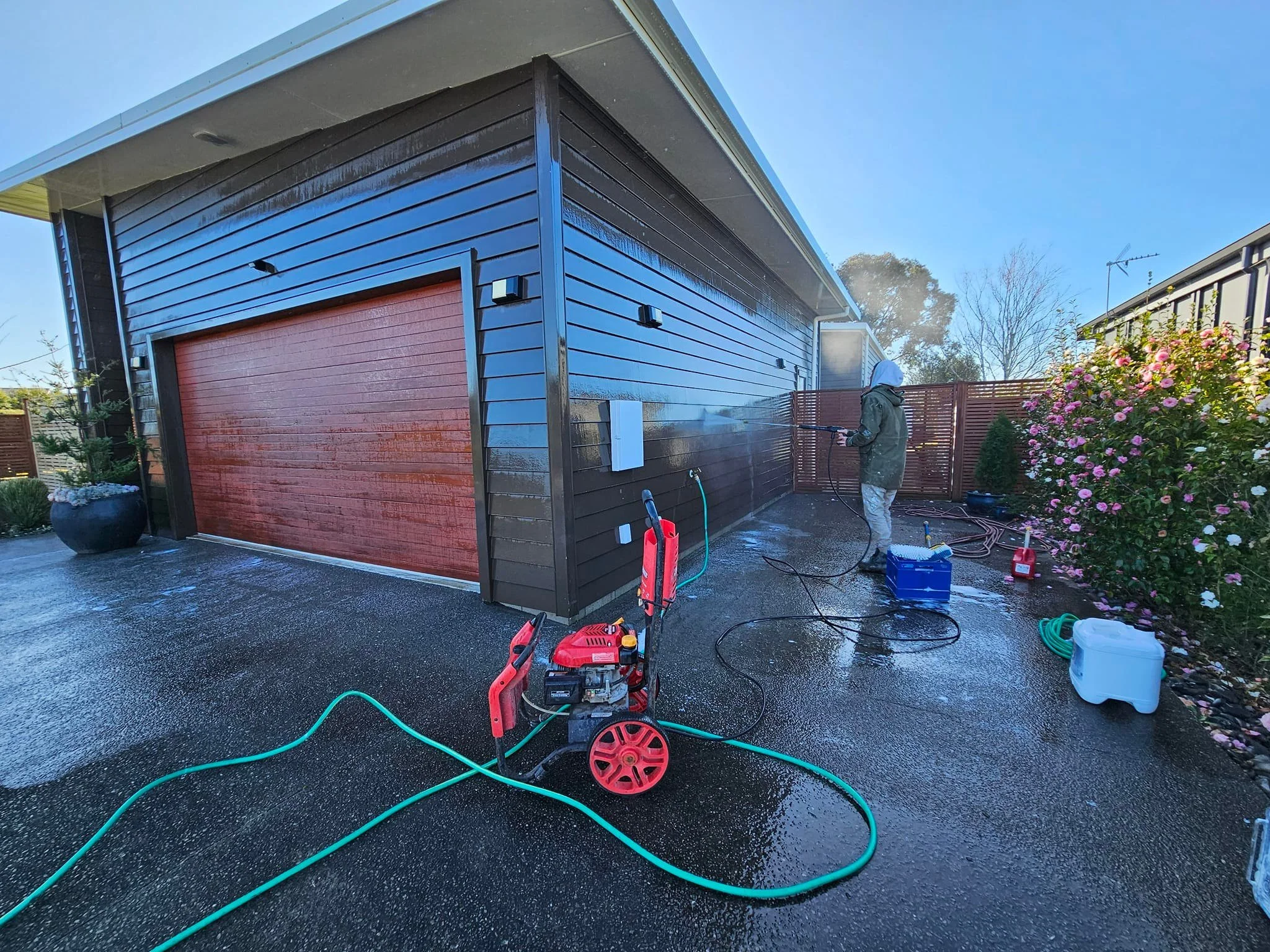 Person washing the side of a house with a pressure washer, with gardening tools and hoses nearby.