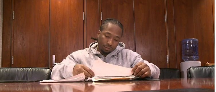 Man reading papers at a conference table in a room with wooden cabinets and a water cooler in the background.