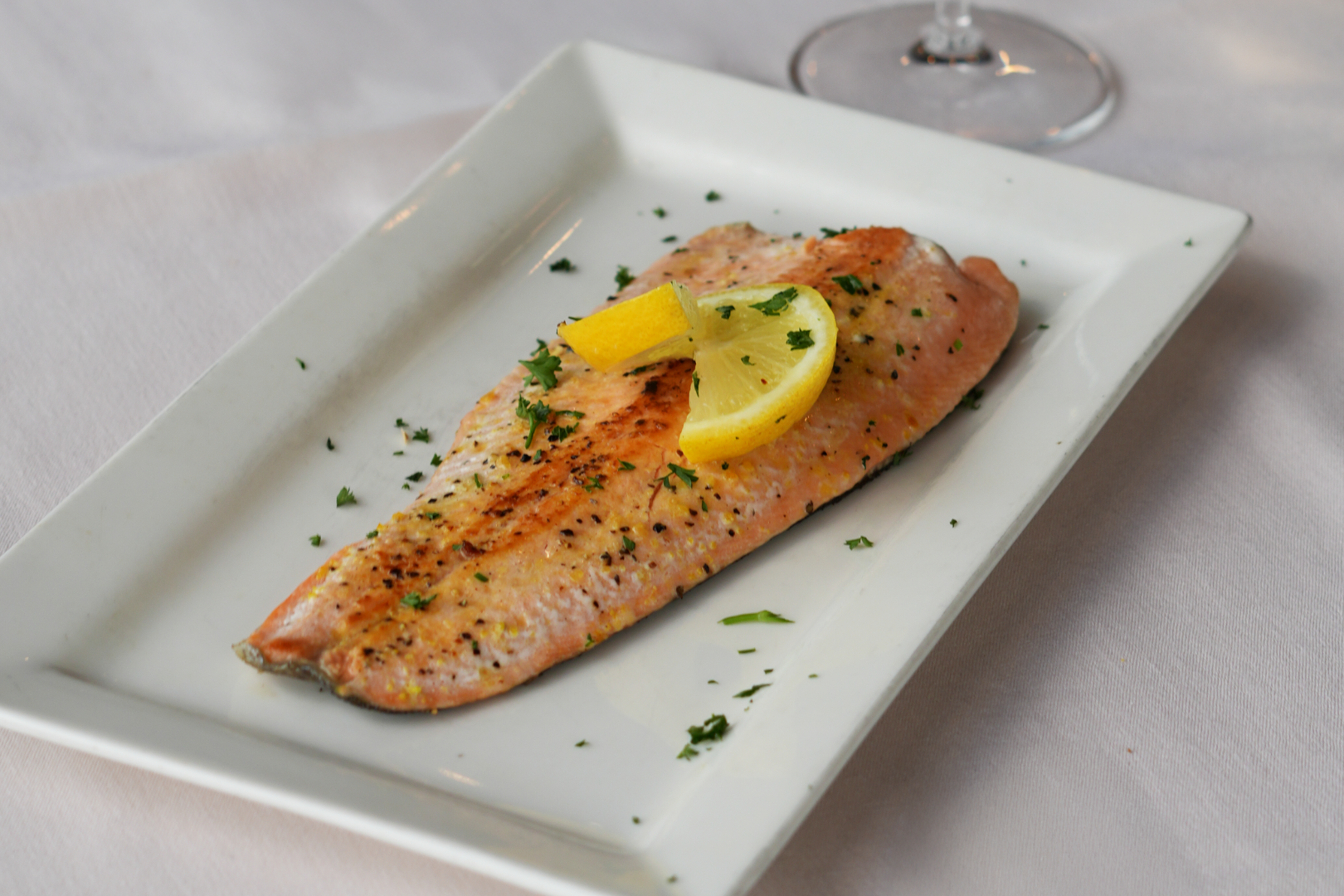 Grilled salmon fillet garnished with lemon wedge and chopped parsley, served on a rectangular white plate.