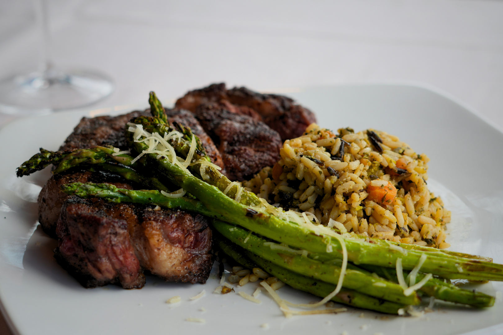 A white plate with grilled steak, roasted asparagus topped with grated cheese, and a serving of rice with vegetables.