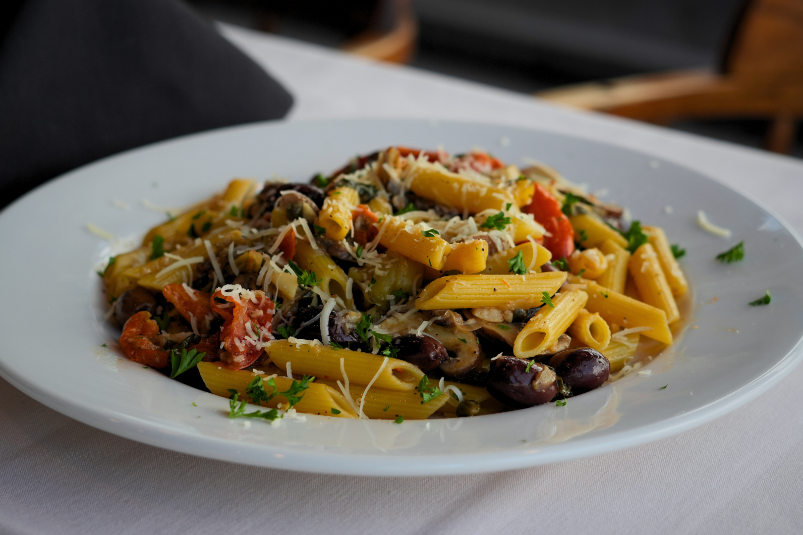 A plate of cooked pasta with black olives, chopped tomatoes, grated cheese, and herbs on a white tablecloth.