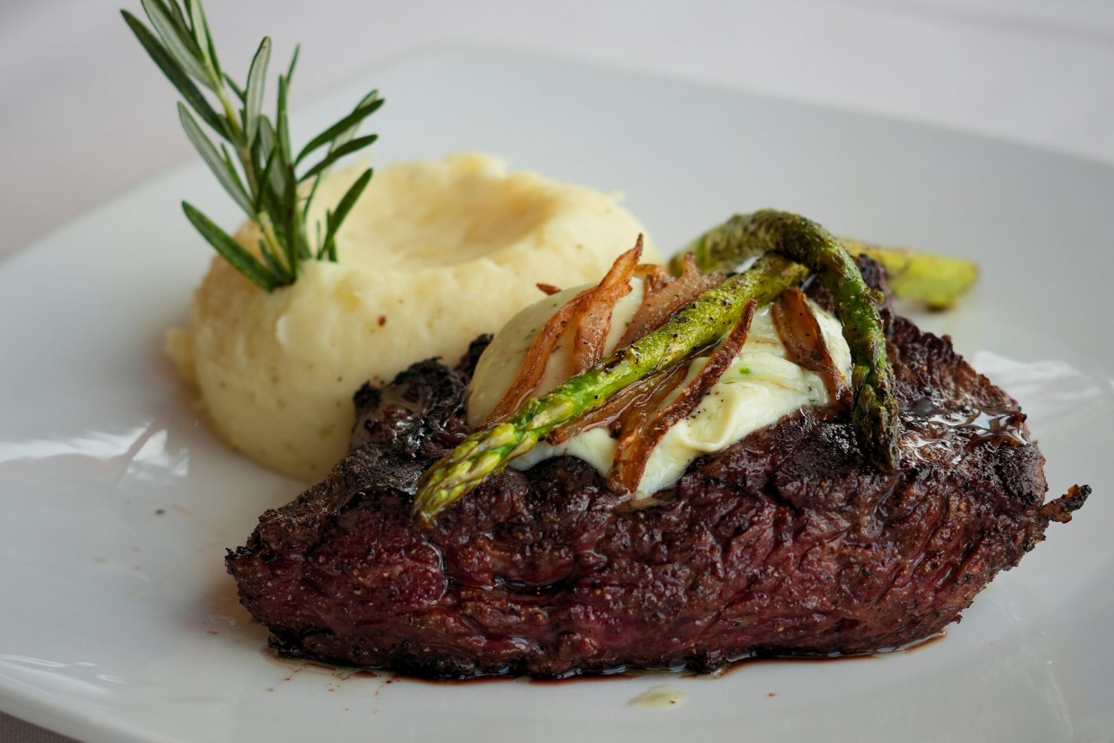 Grilled steak topped with fried onions and green asparagus, served with mashed potatoes garnished with a sprig of rosemary.