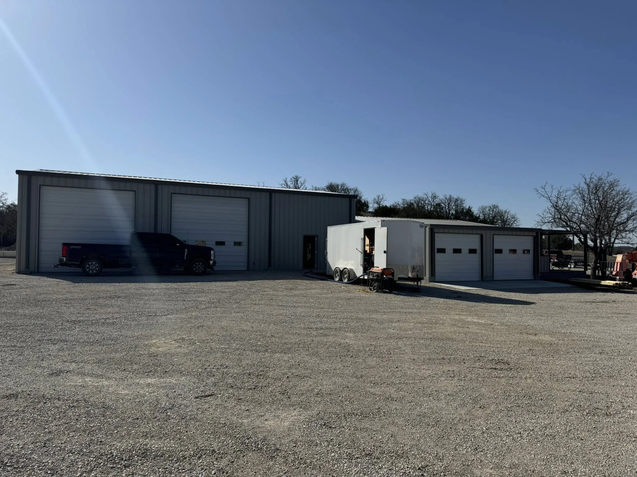 Large metal building with multiple garage doors, white enclosed trailer, black pickup truck parked nearby, and gravel lot under clear blue sky.