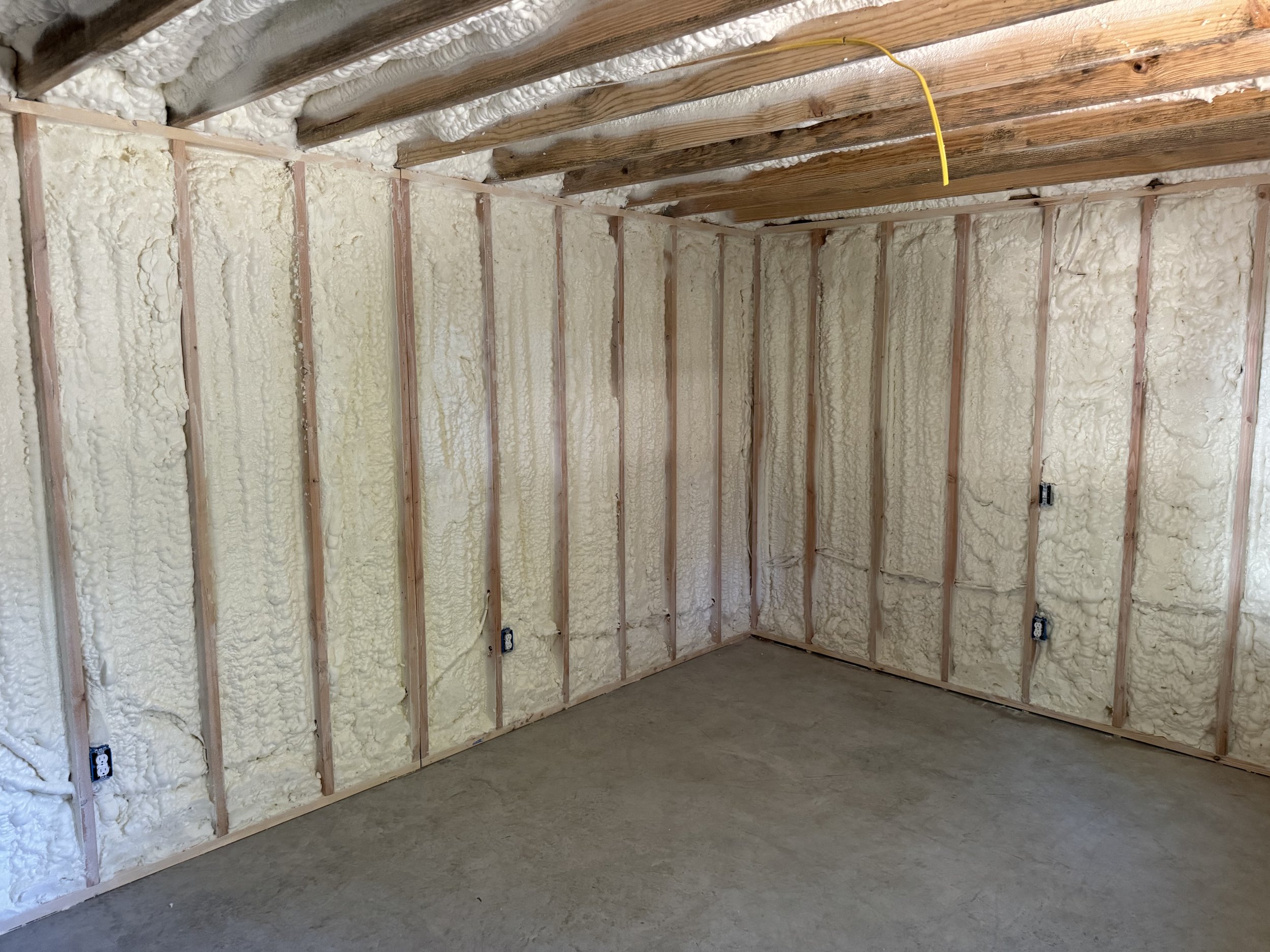 Unfinished room with foam insulation on the walls, exposed wooden beams on the ceiling, electrical outlets and wiring visible, and a concrete floor.