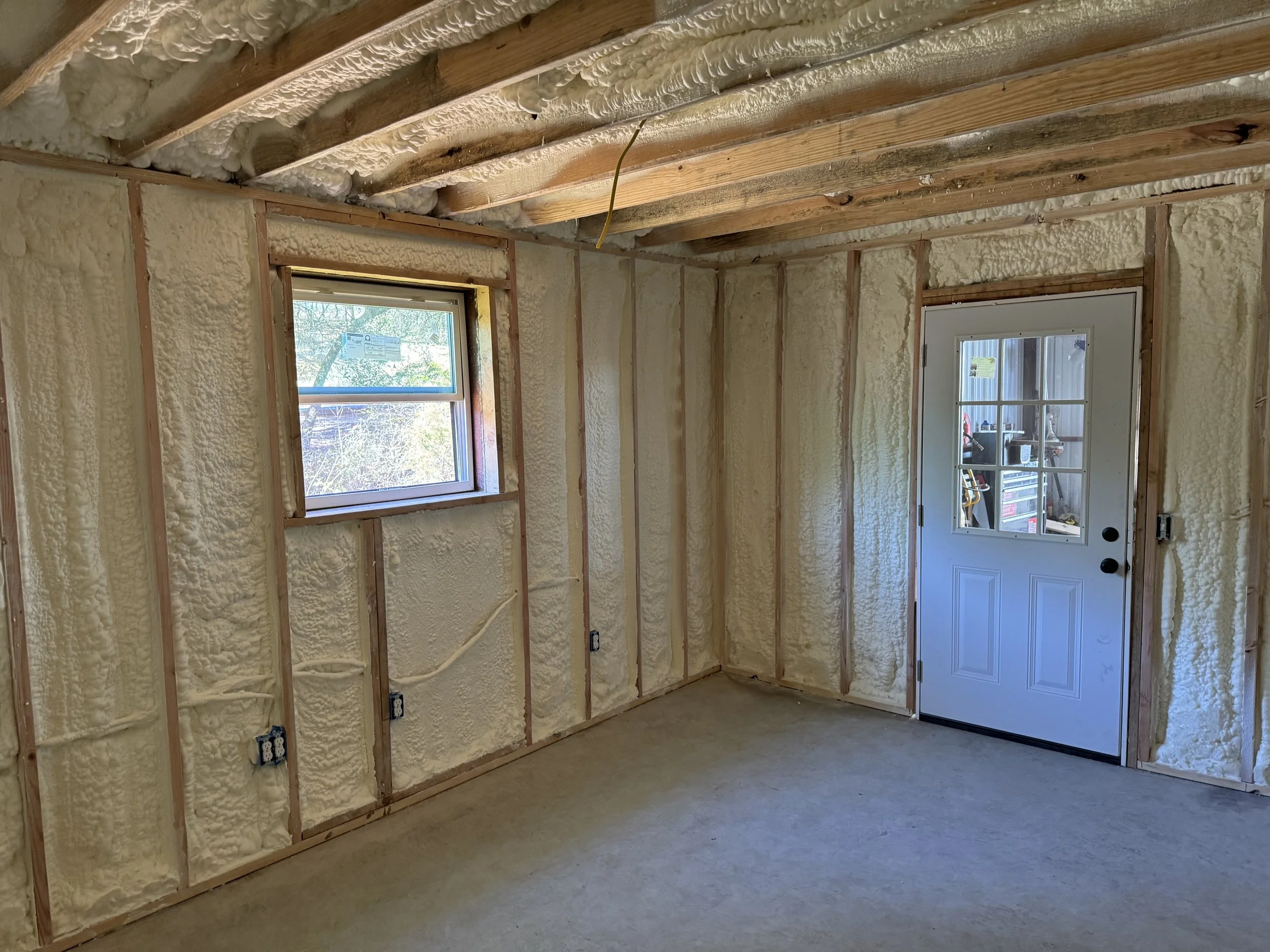 Interior of a room under construction with exposed wooden studs and insulation, a window showing outdoor trees, and a white door with glass panes.