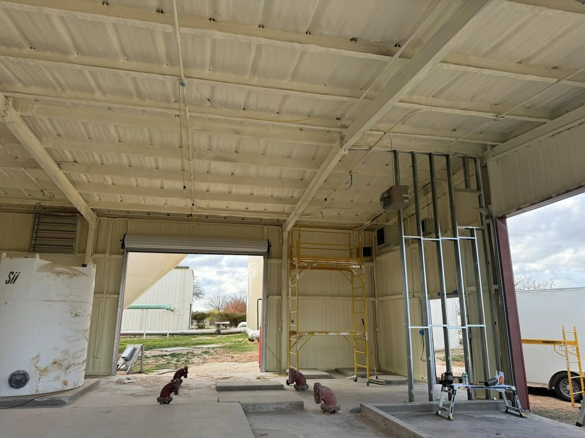 Interior of a construction site showing metal framing, yellow scaffolding, a partially open roller door, and construction tools, with an outdoor view of pipes and trees.