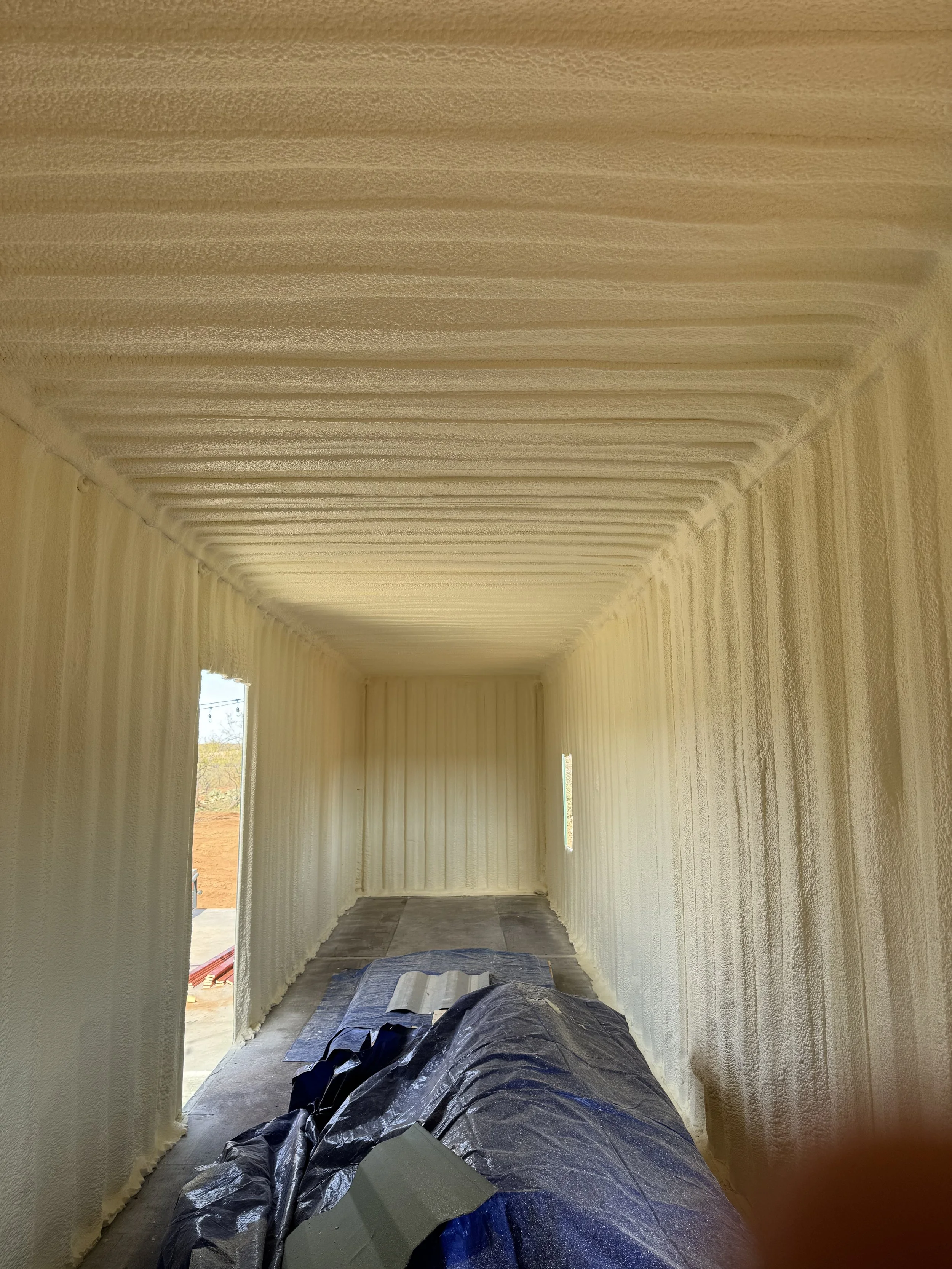 Interior of a narrow room with walls and ceiling covered in spray foam insulation. There are two open doorways on the left side and a small window on the right side, with construction materials and a plastic sheet on the floor.