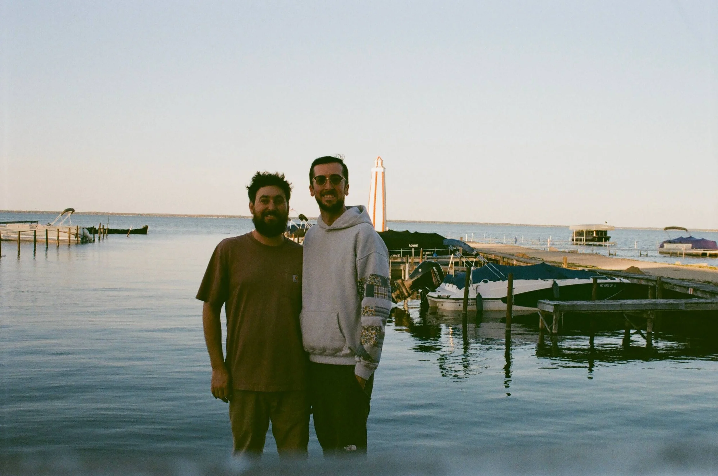 Two men standing by the water at a marina with boats and a lighthouse in the background during sunset.