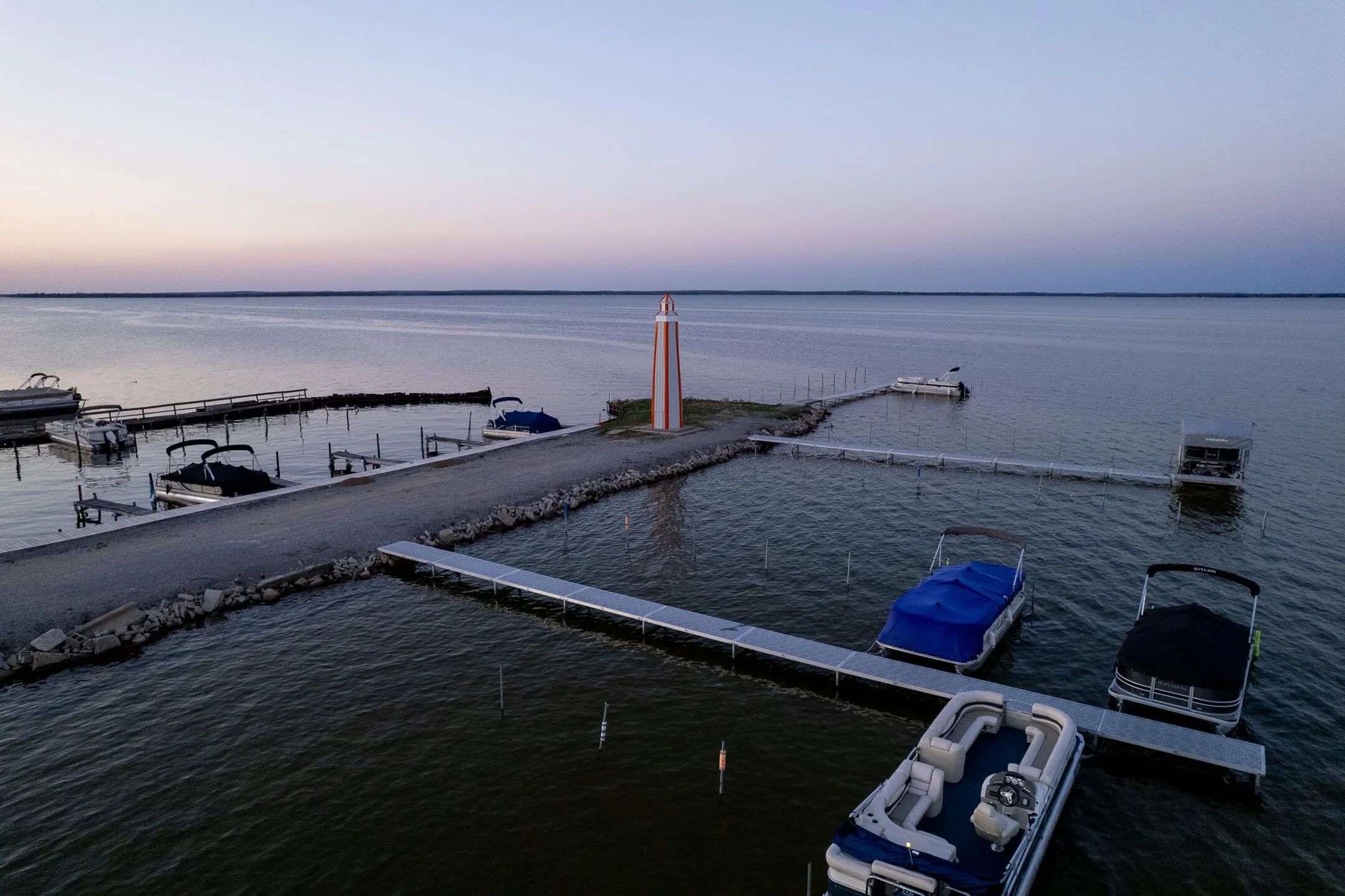 A marina at dusk with several boats docked, a narrow pier, and a red and white navigation marker in the middle of the water.
