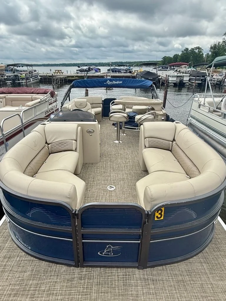 A pontoon boat with beige leather seating, a small table, and a steering console, docked at a marina with other boats and cloudy skies in the background.