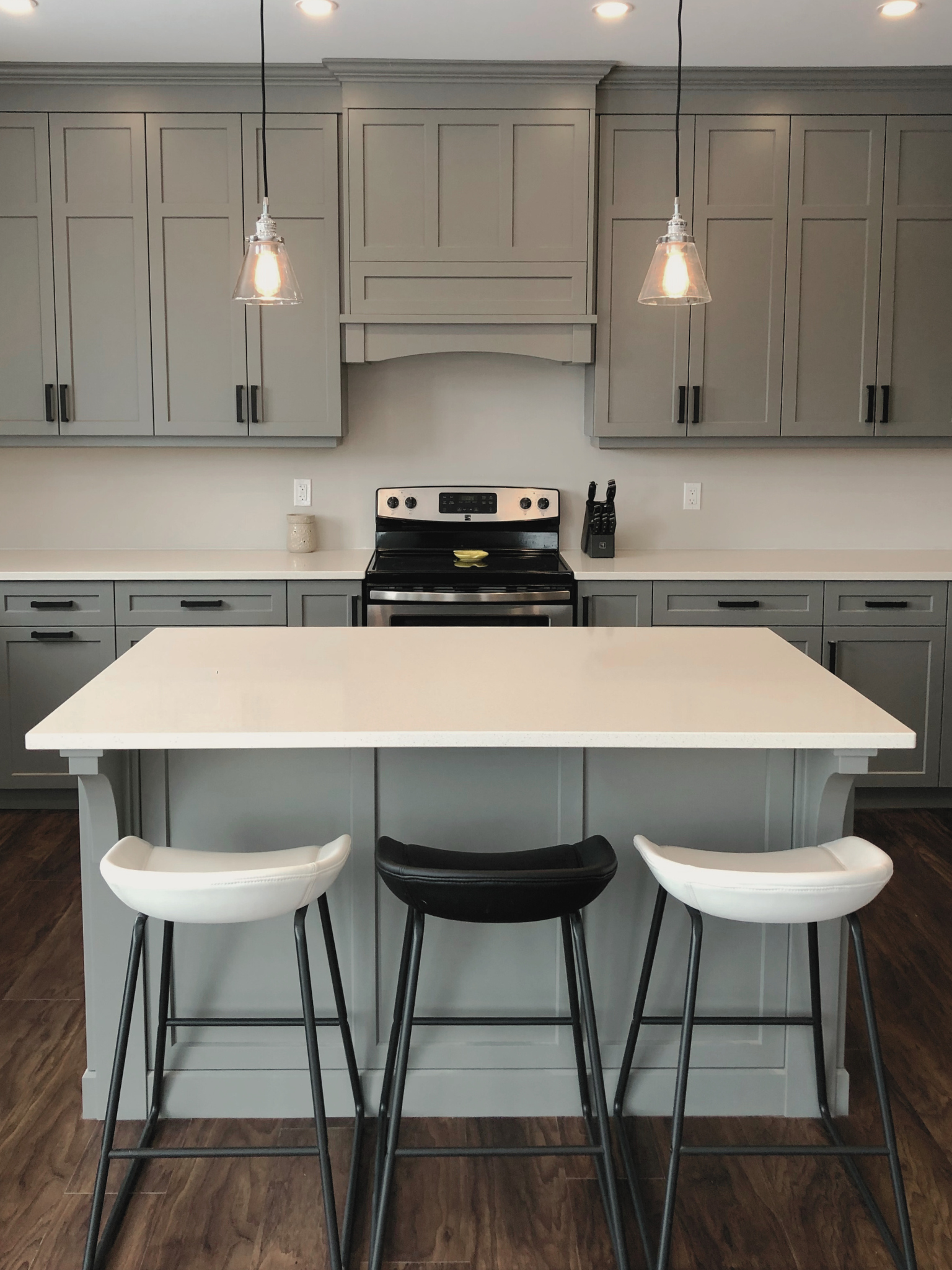 Modern kitchen with light gray cabinets, white countertop, black and white barstools, and stainless steel stove. There are pendant lights hanging above the island.