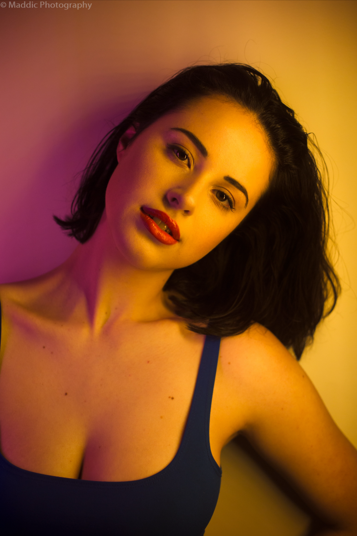 A woman with dark hair and red lipstick looking at the camera, wearing a blue top, illuminated with yellow and pink lighting.