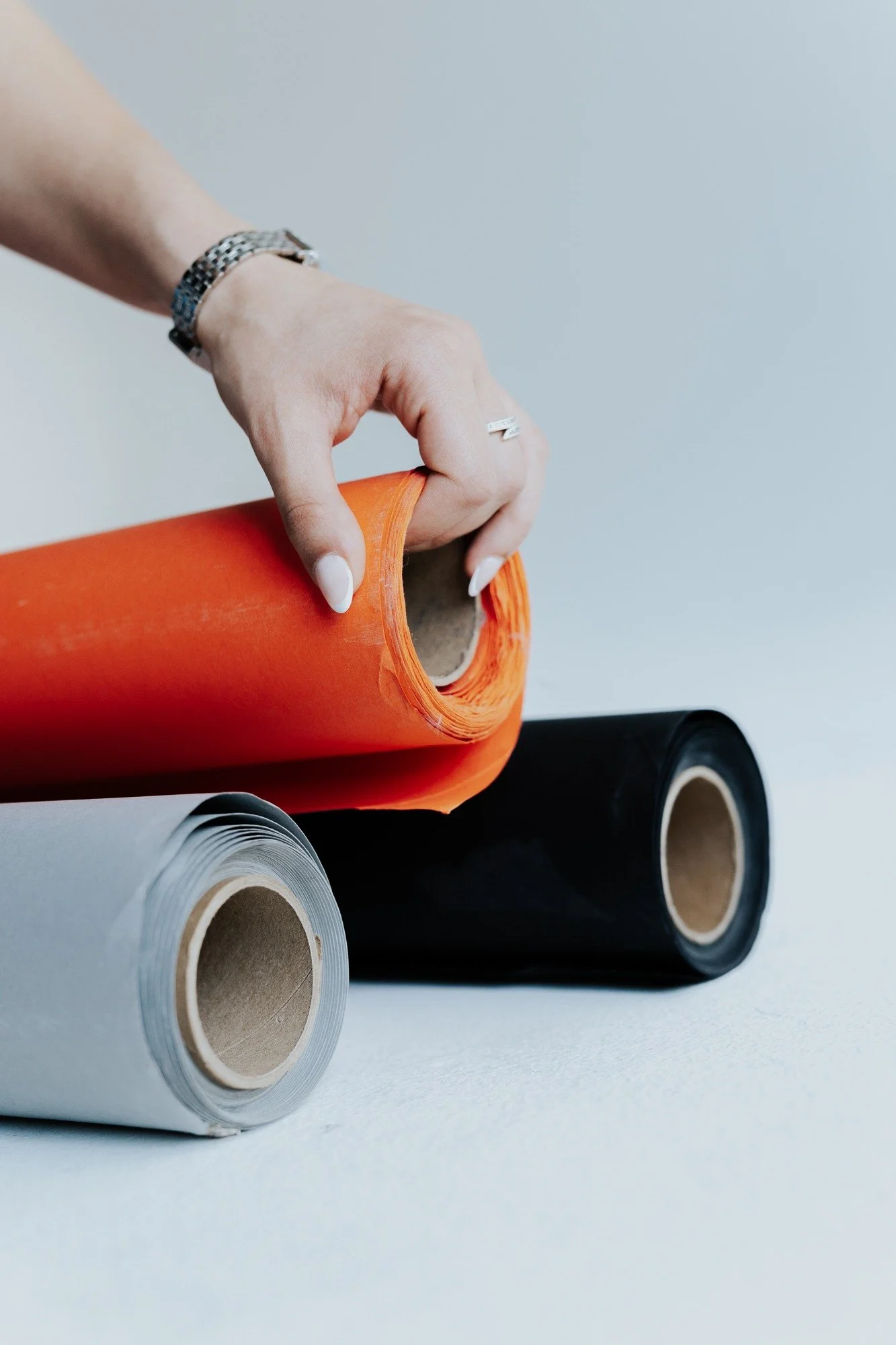 Person's hand holding a roll of orange vinyl sheet over two rolls of black and gray vinyl sheets on a light surface.