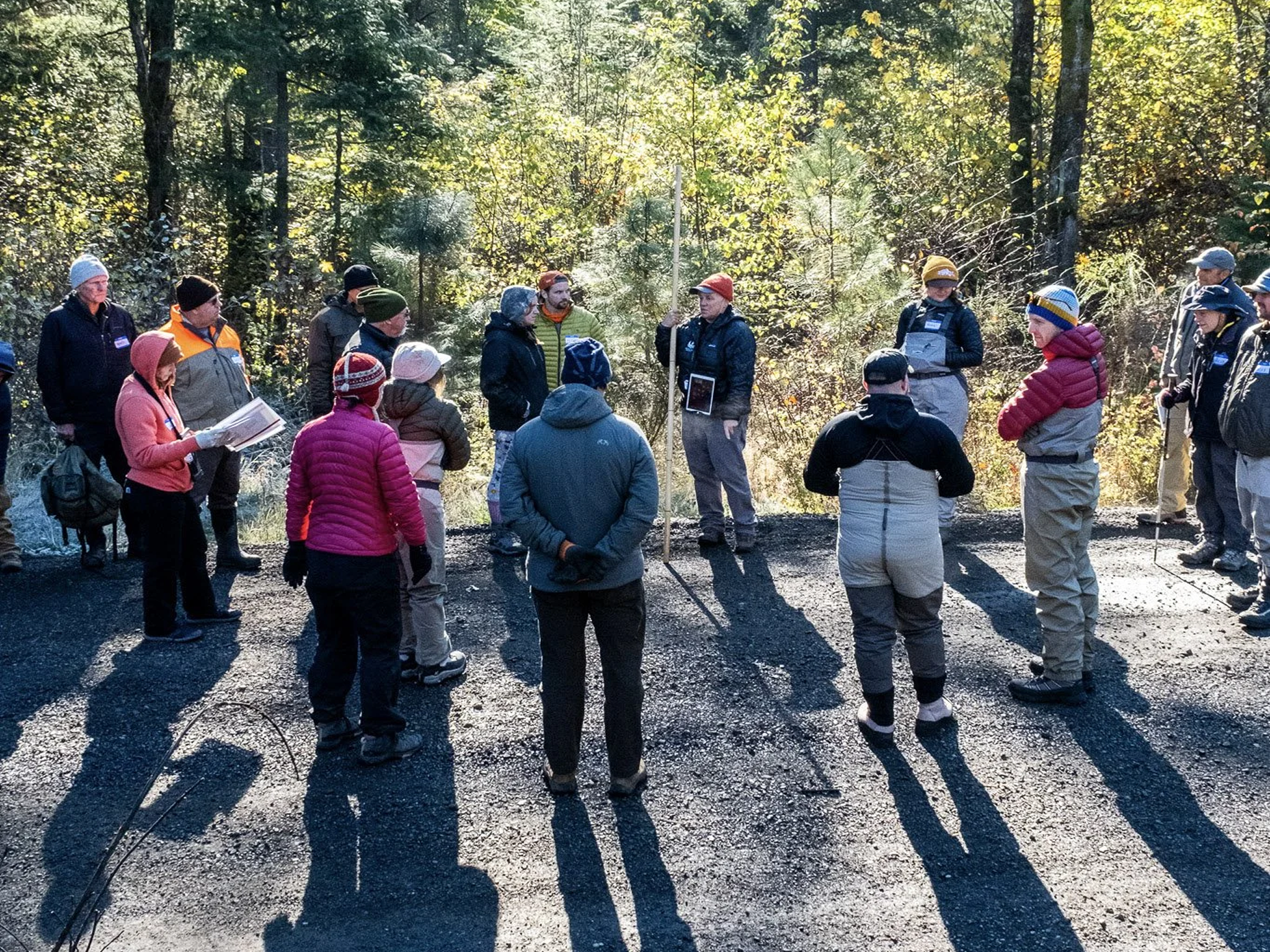 Group of people gathered outdoors in a forested area, listening to a guide or speaker, dressed in outdoor winter clothing and holding a measuring stick.
