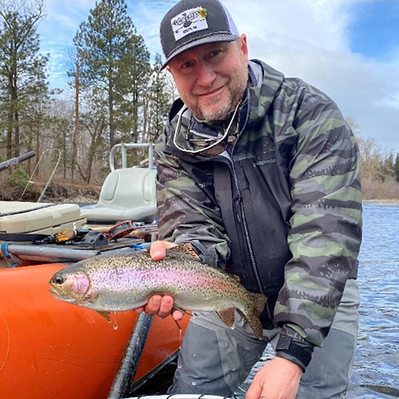 Man smiling and holding a rainbow trout fish he caught while fishing on a river, with trees and fishing equipment visible in the background.