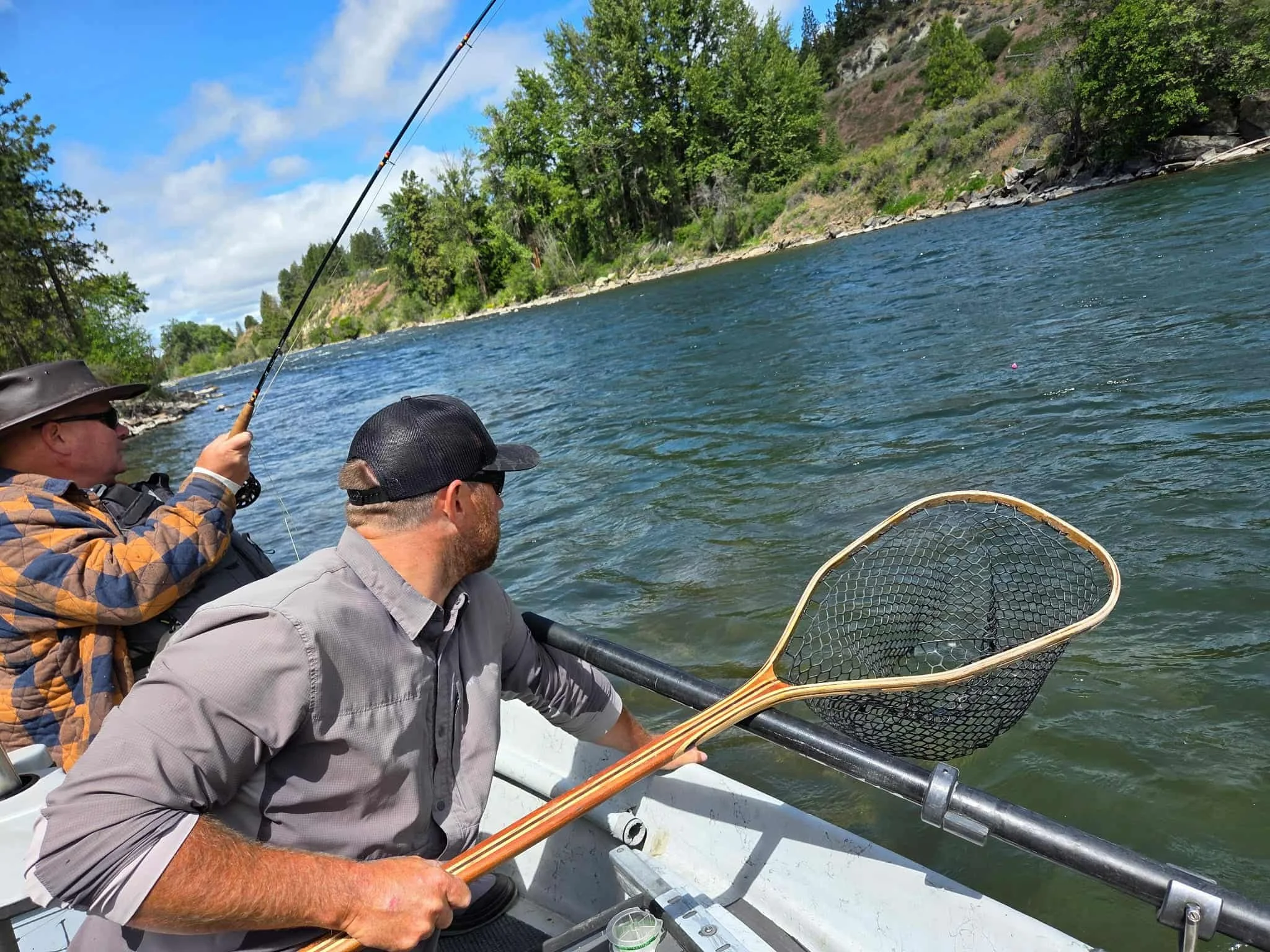 Two men fishing from a boat on a river, one holding a fishing rod and the other holding a net, with lush green trees and rocky hills in the background.