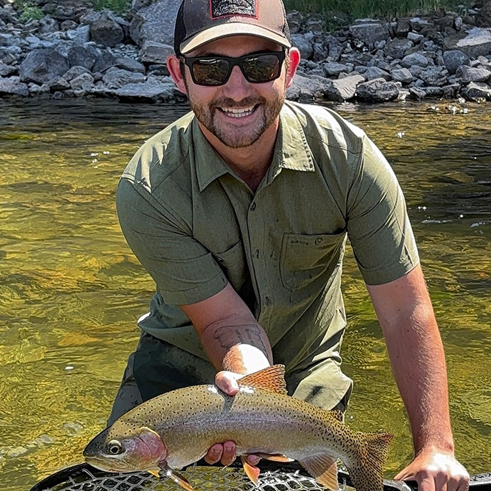 A man smiling while holding a large fish he caught in a river with rocky banks.