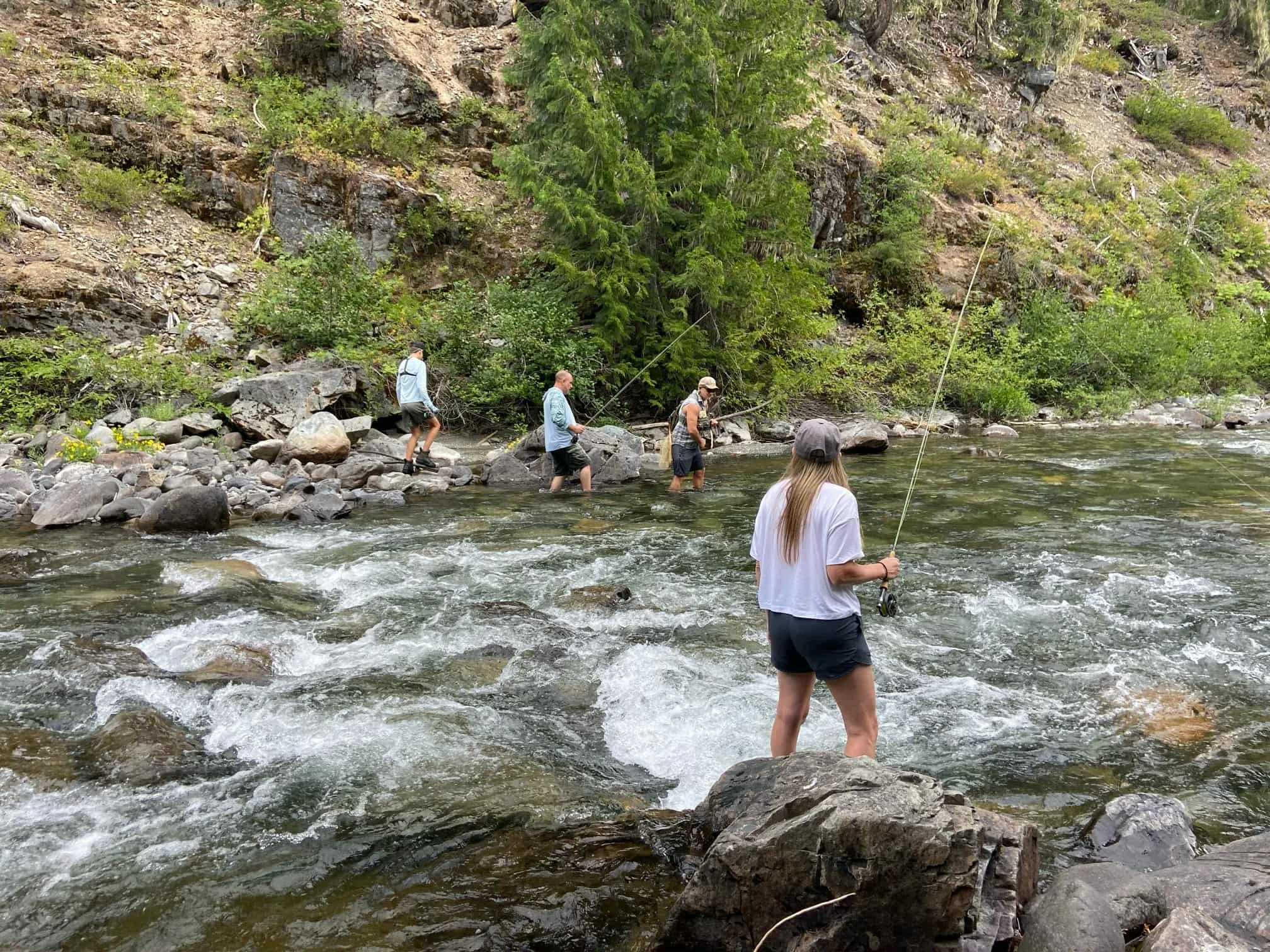 People fishing in a river surrounded by rocks, trees, and a hillside.