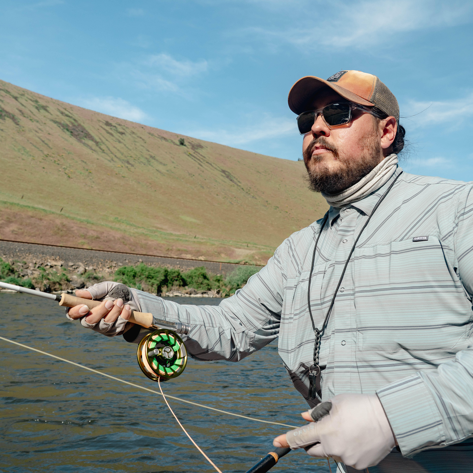 Man fishing in a river with hills in the background, wearing sunglasses, a cap, a striped long-sleeve shirt, gloves, and a neck gaiter.
