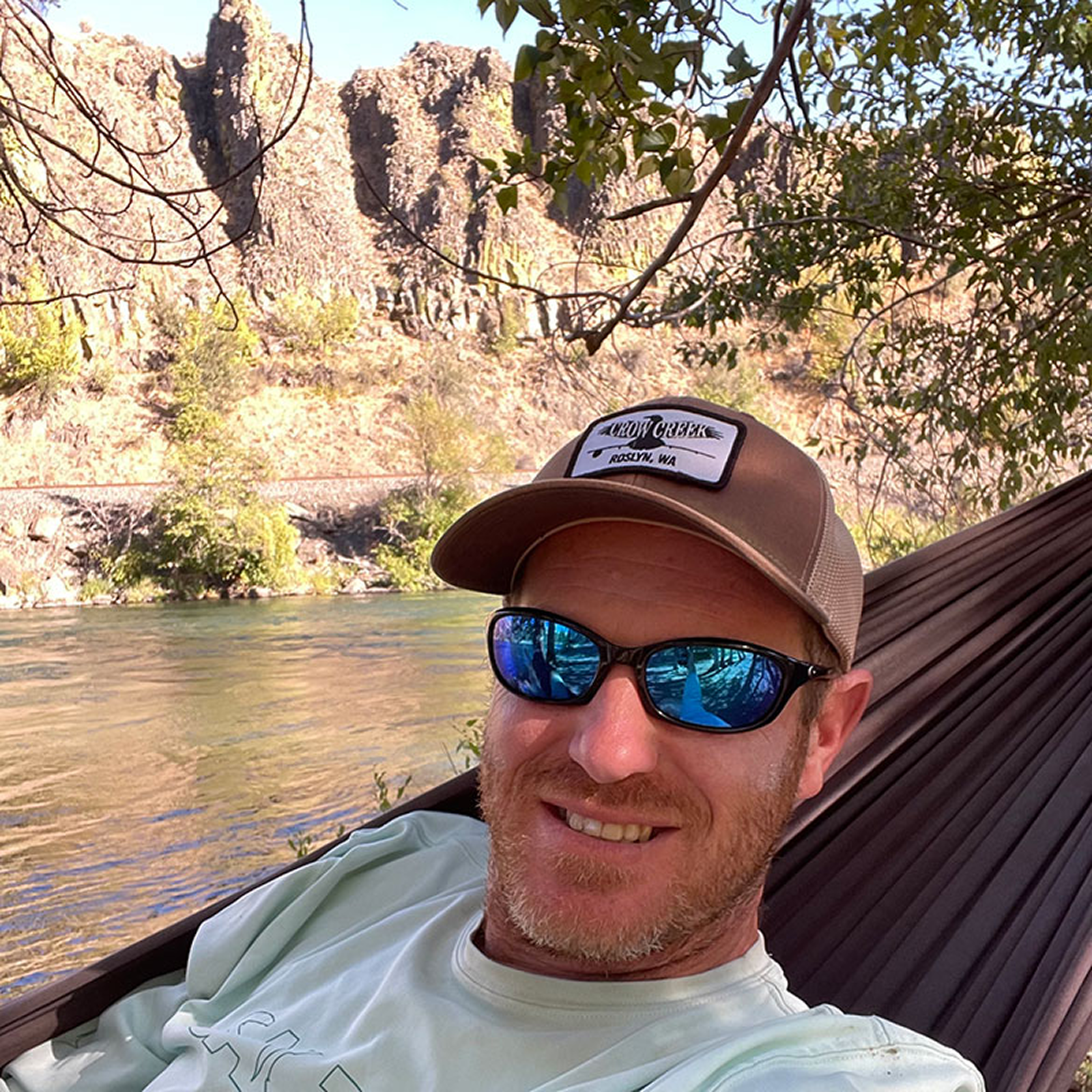 A man with a beard and sunglasses relaxing on a hammock near a river, with rocky cliffs and trees in the background.