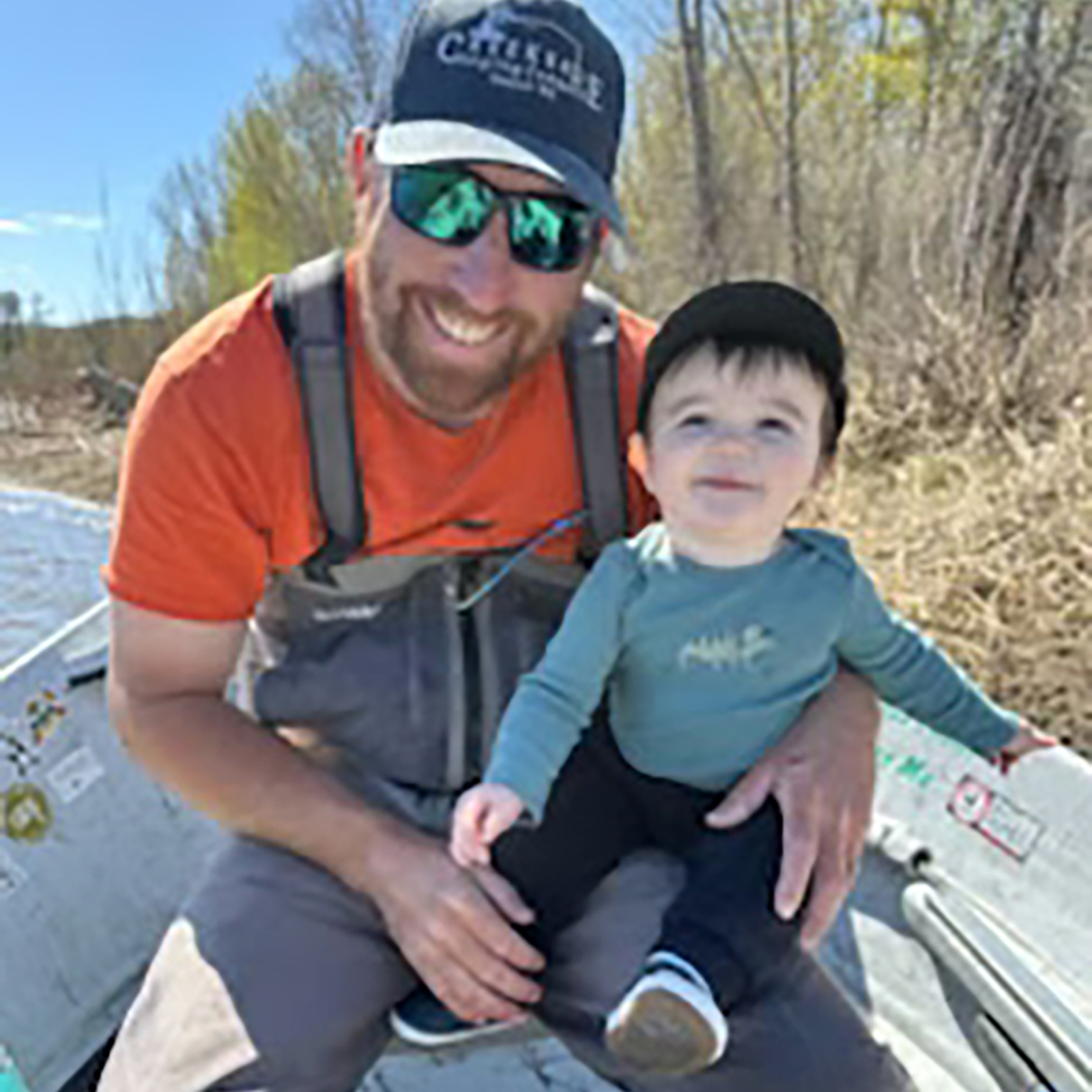 Man in orange shirt and sunglasses holding smiling young child in outdoor setting with trees