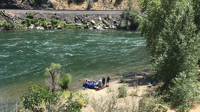 A group of people on a beach by a river, with a blue inflatable boat and lush green trees surrounding the area.